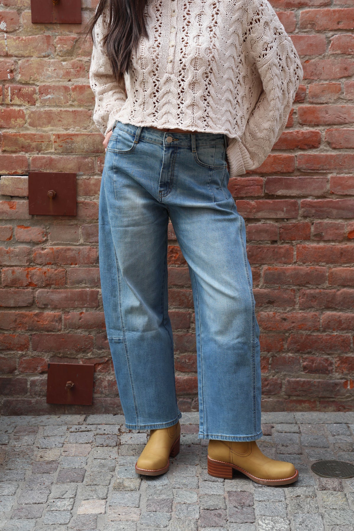Woman wearing barrel jeans with a cream knit against a brick wall.