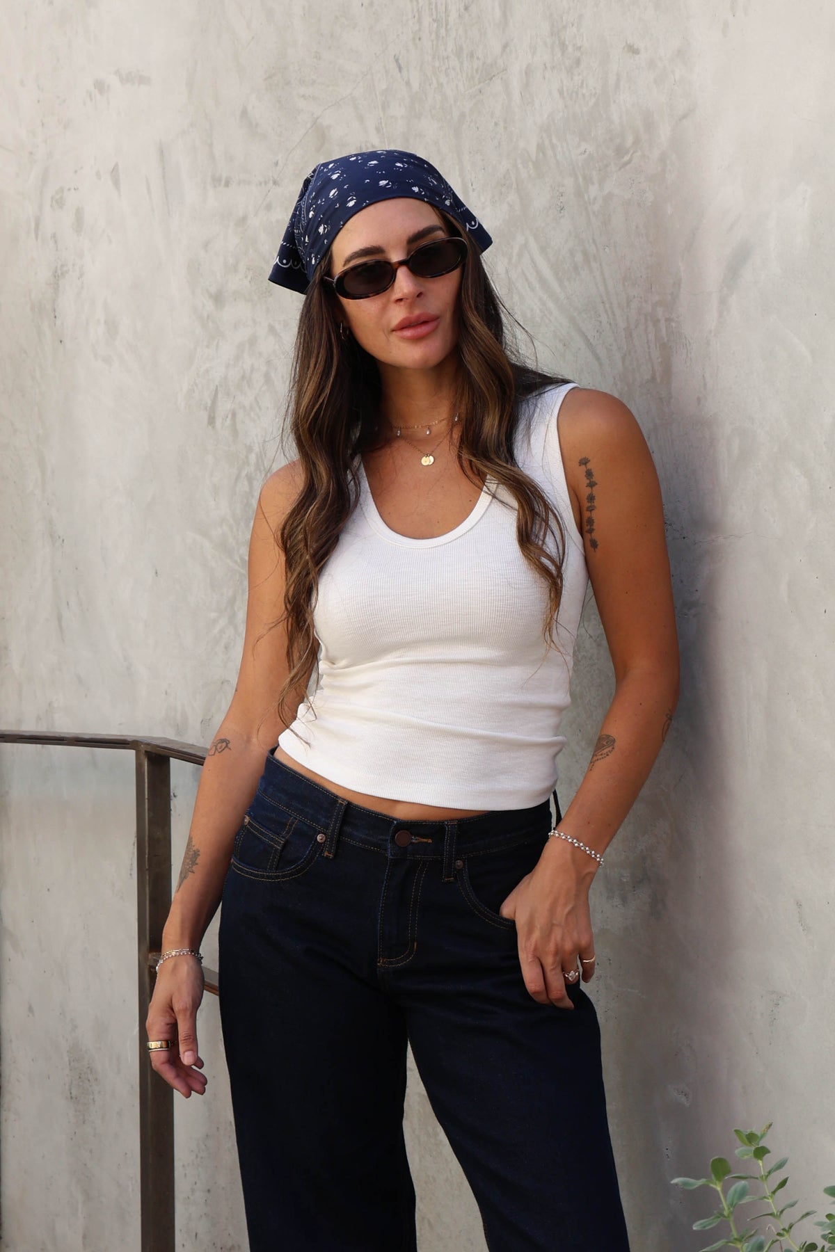 Woman wearing a white top, jeans and a bandana against a plain background.