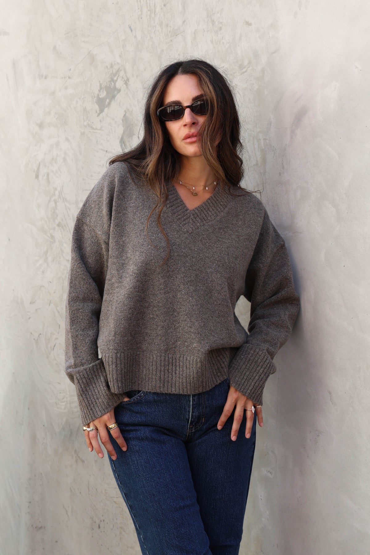 Woman wearing a grey v-neck sweater and jeans against a plain background.