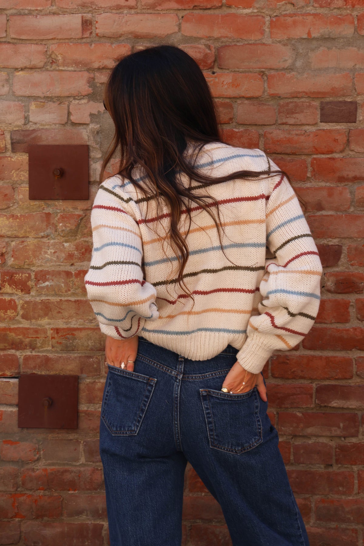 Woman wearing a colorful striped sweater with jeans against a brick background.