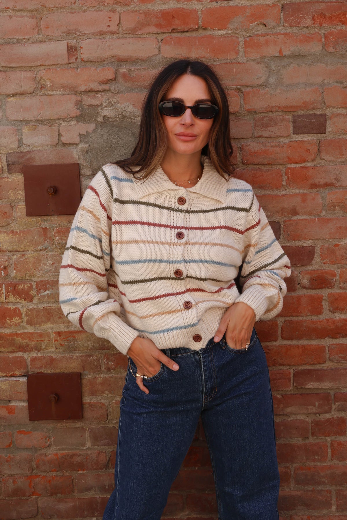 Woman wearing a colorful striped sweater with jeans against a brick background.