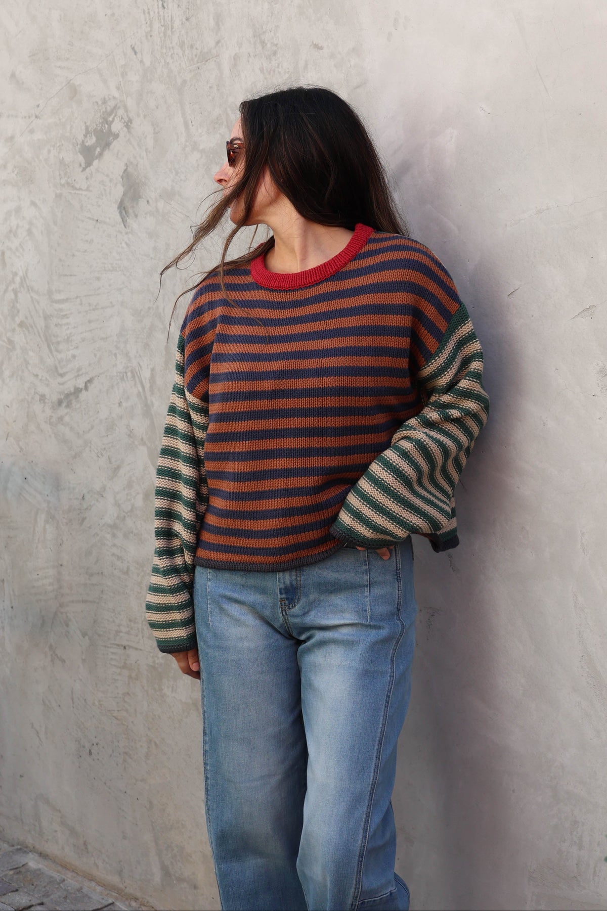 A woman wearing a striped knit sweater with a red crew neck and blue jeans, leaning against a textured gray concrete wall.