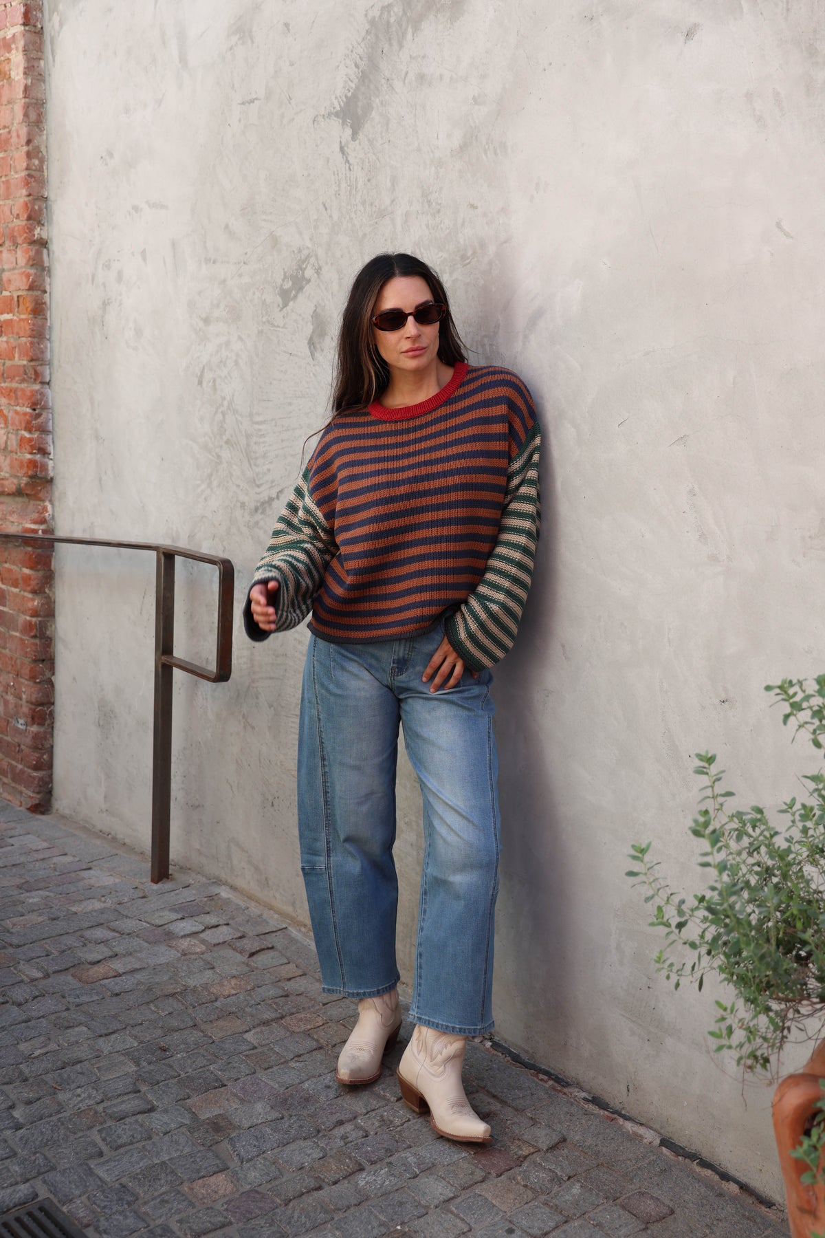 A woman wearing a striped knit sweater with a red collar and blue jeans, leaning against a textured gray concrete wall.
