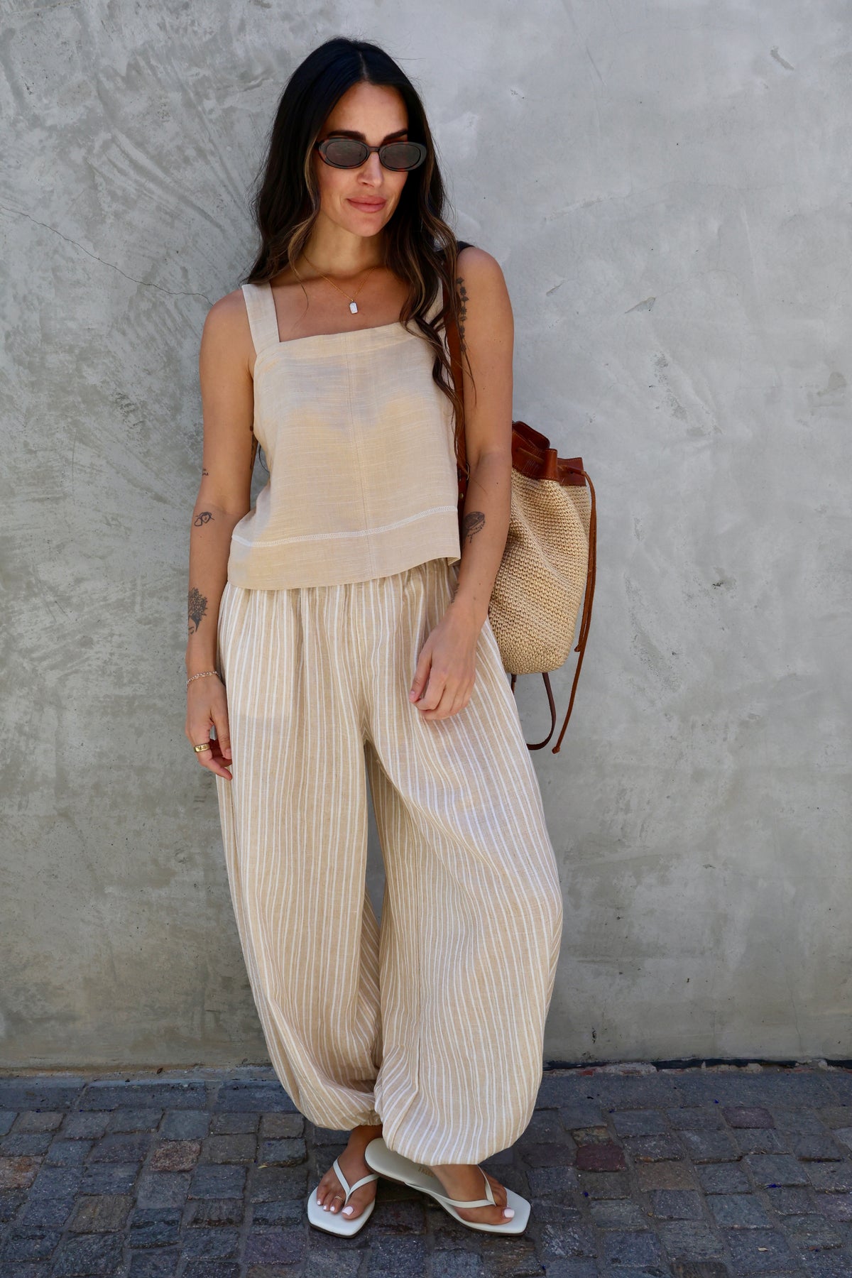 A woman stands against a textured gray wall wearing a beige sleeveless top and matching pleated wide-leg pants, with sunglasses, a woven straw bag, and flat sandals.