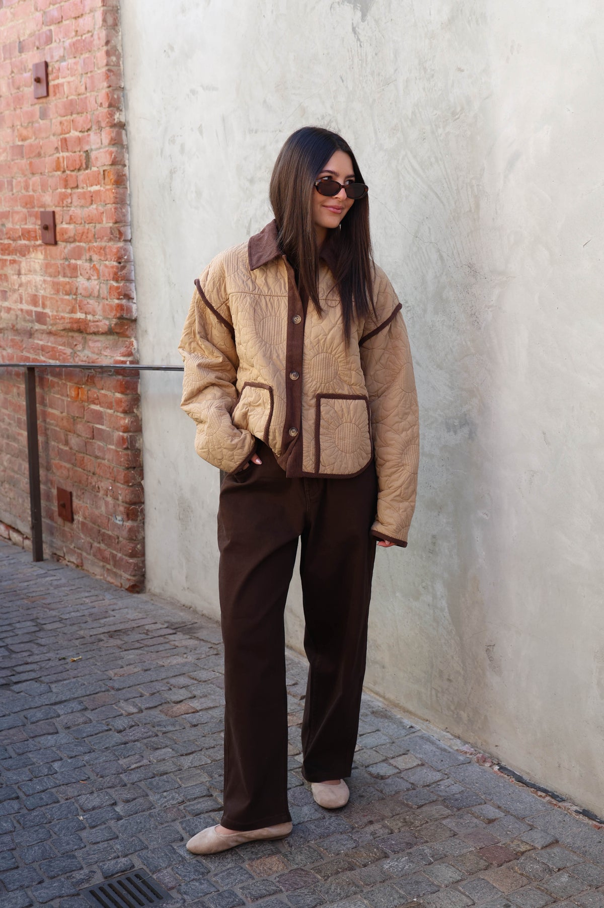 Woman wearing a brown jacket, brown pants holding a brown bad in front of a plain wall.