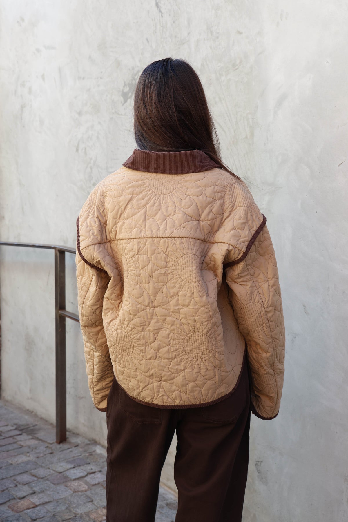 Woman wearing a brown jacket, brown pants holding a brown bad in front of a plain wall.