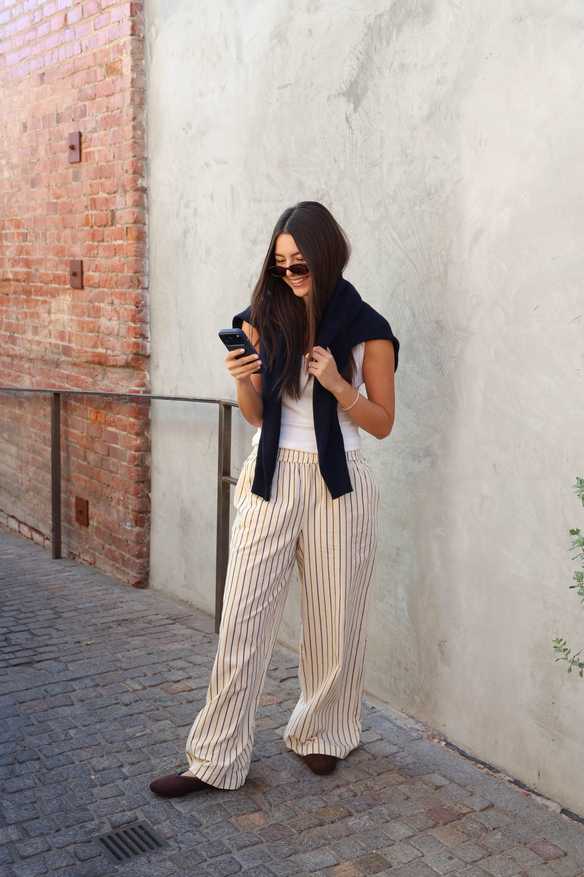 Woman wearing a white tank, striped pants in front of a plain wall.