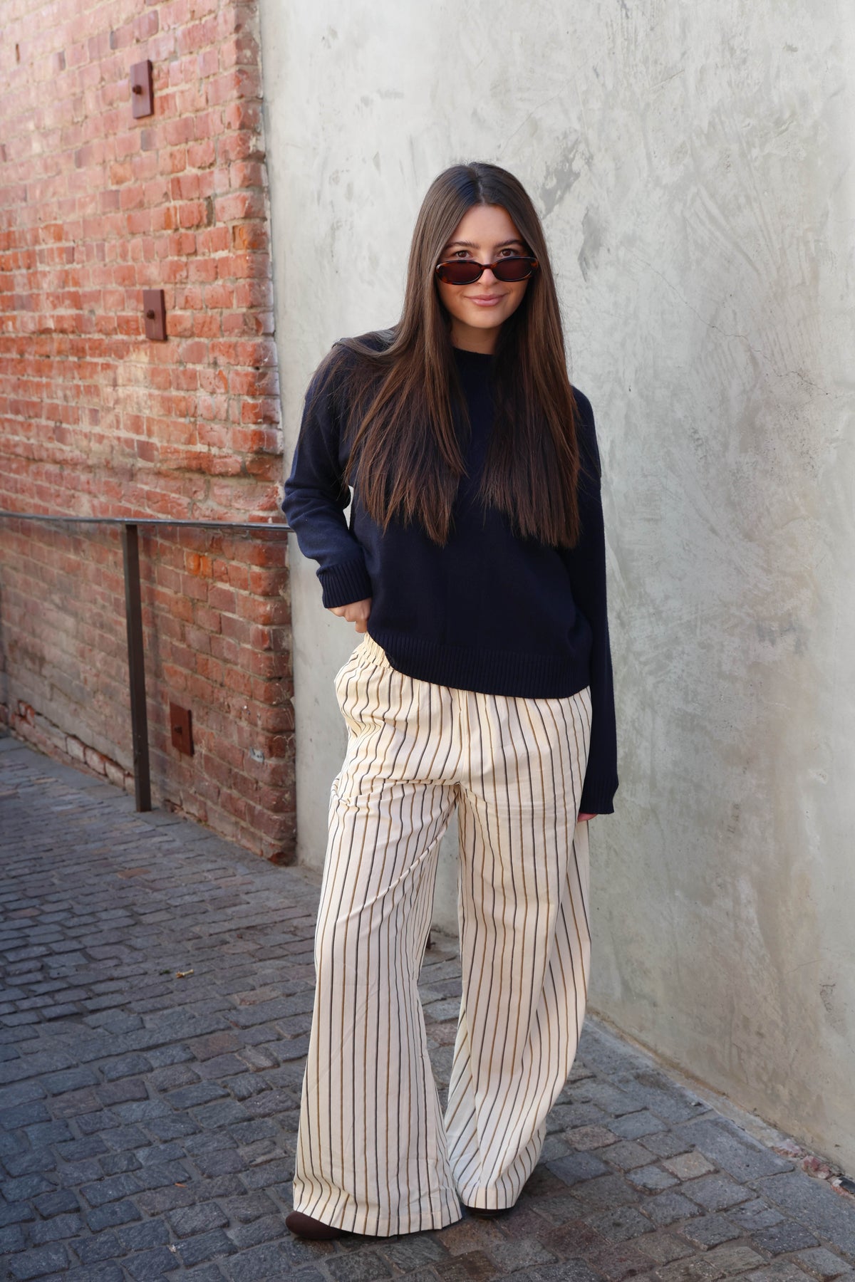Woman wearing a white tank, striped pants in front of a plain wall.