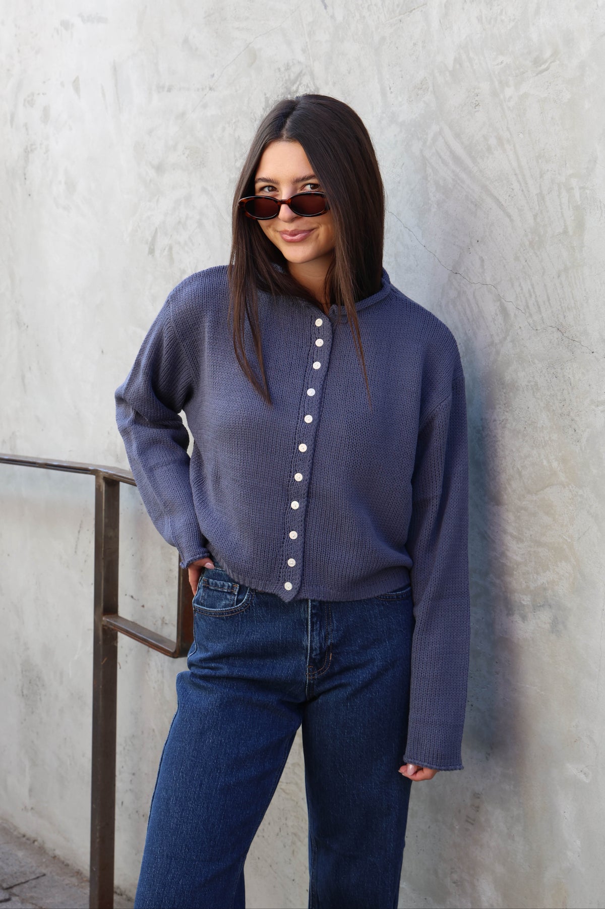 Woman wearing jeans and a blue sweater with buttons standing in front of a plain wall.
