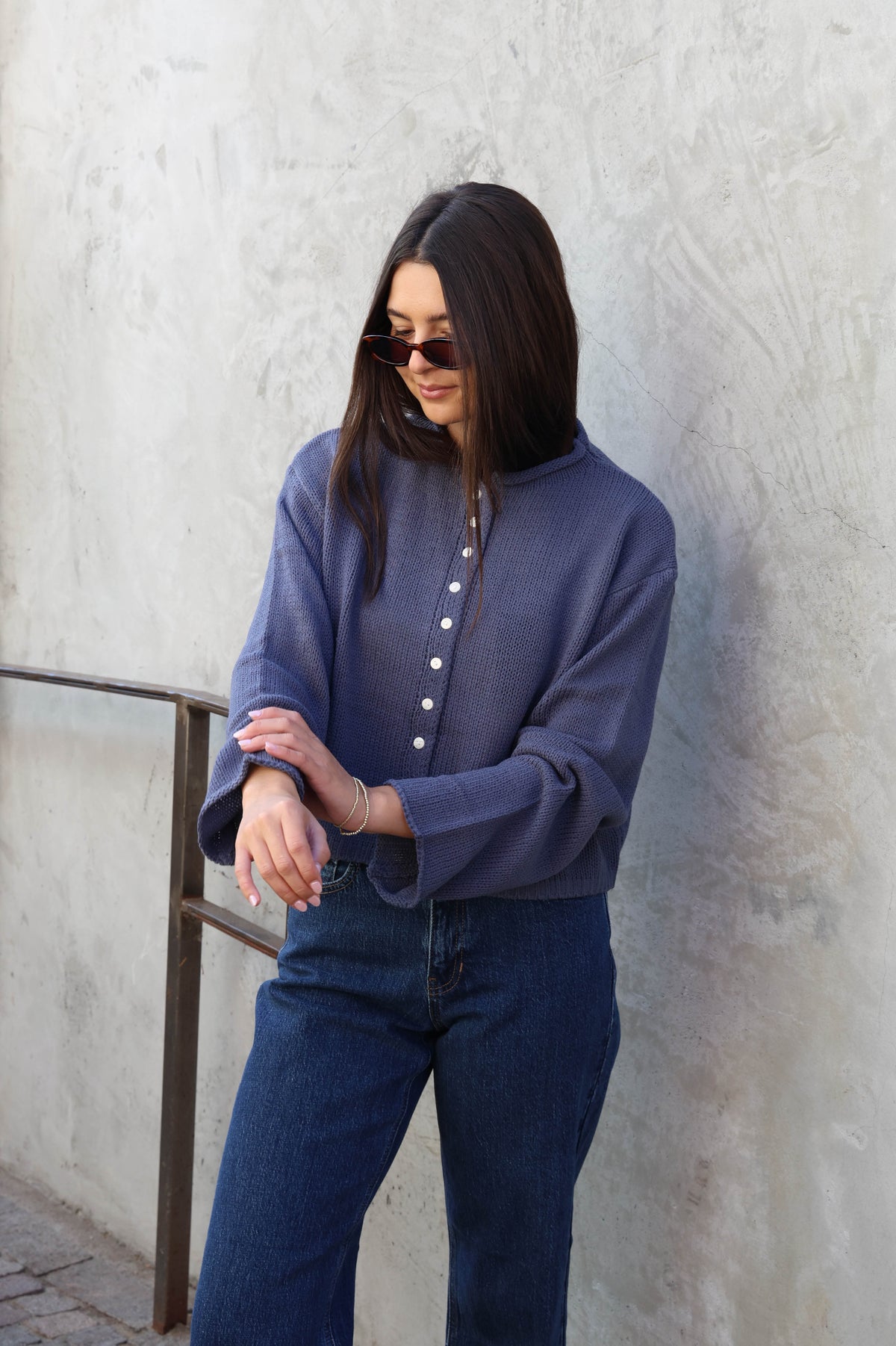Woman wearing jeans and a blue sweater with buttons standing in front of a plain wall.
