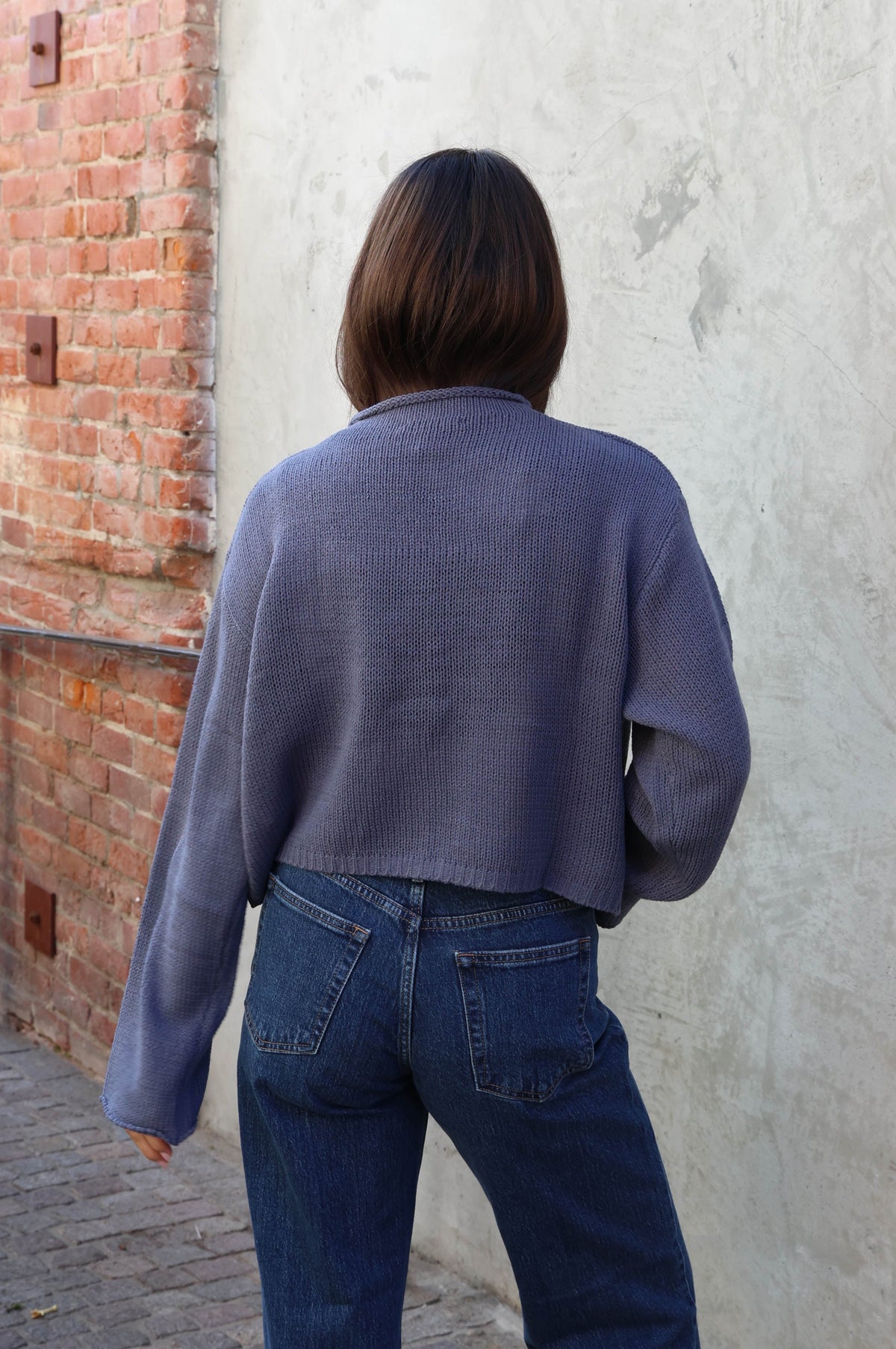 Woman wearing jeans and a blue sweater with buttons standing in front of a plain wall.