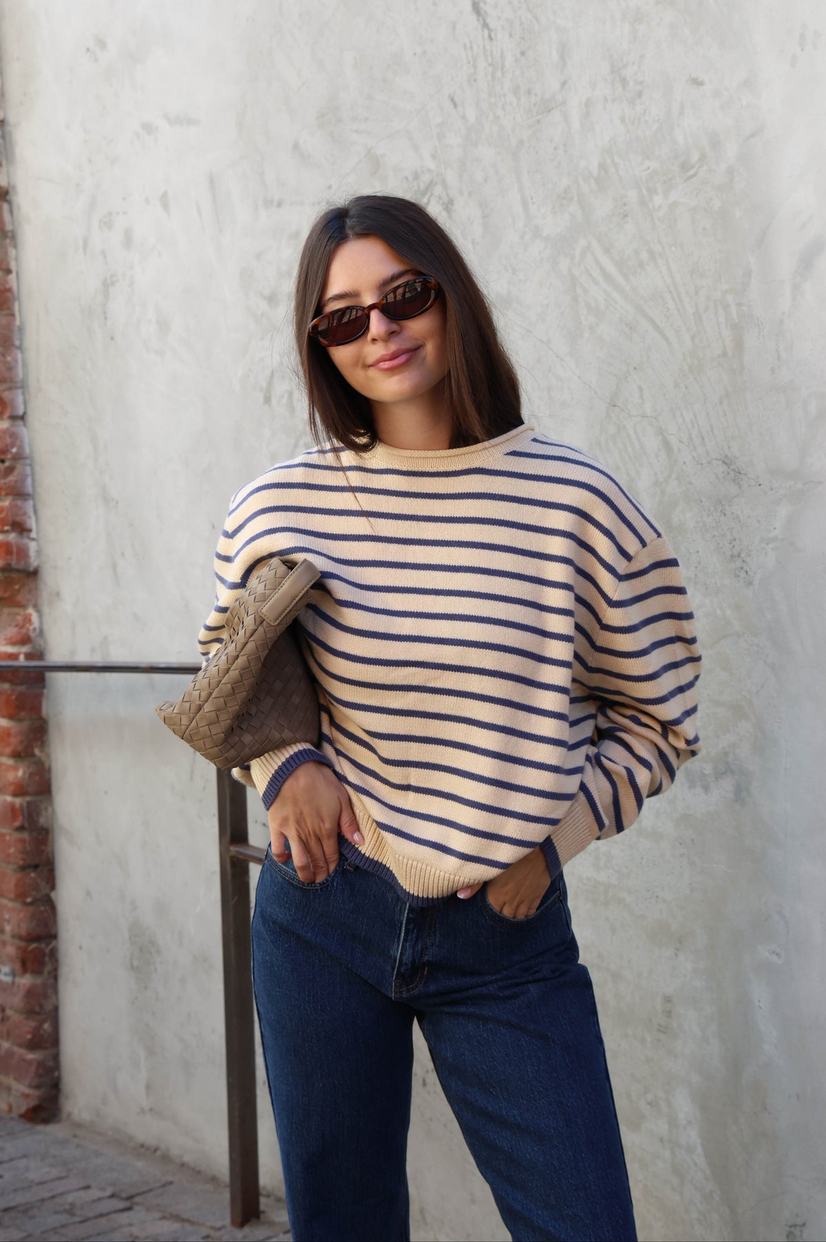 Woman wearing a cream and blue striped sweater with jeans and sunglasses standing against a plain background.