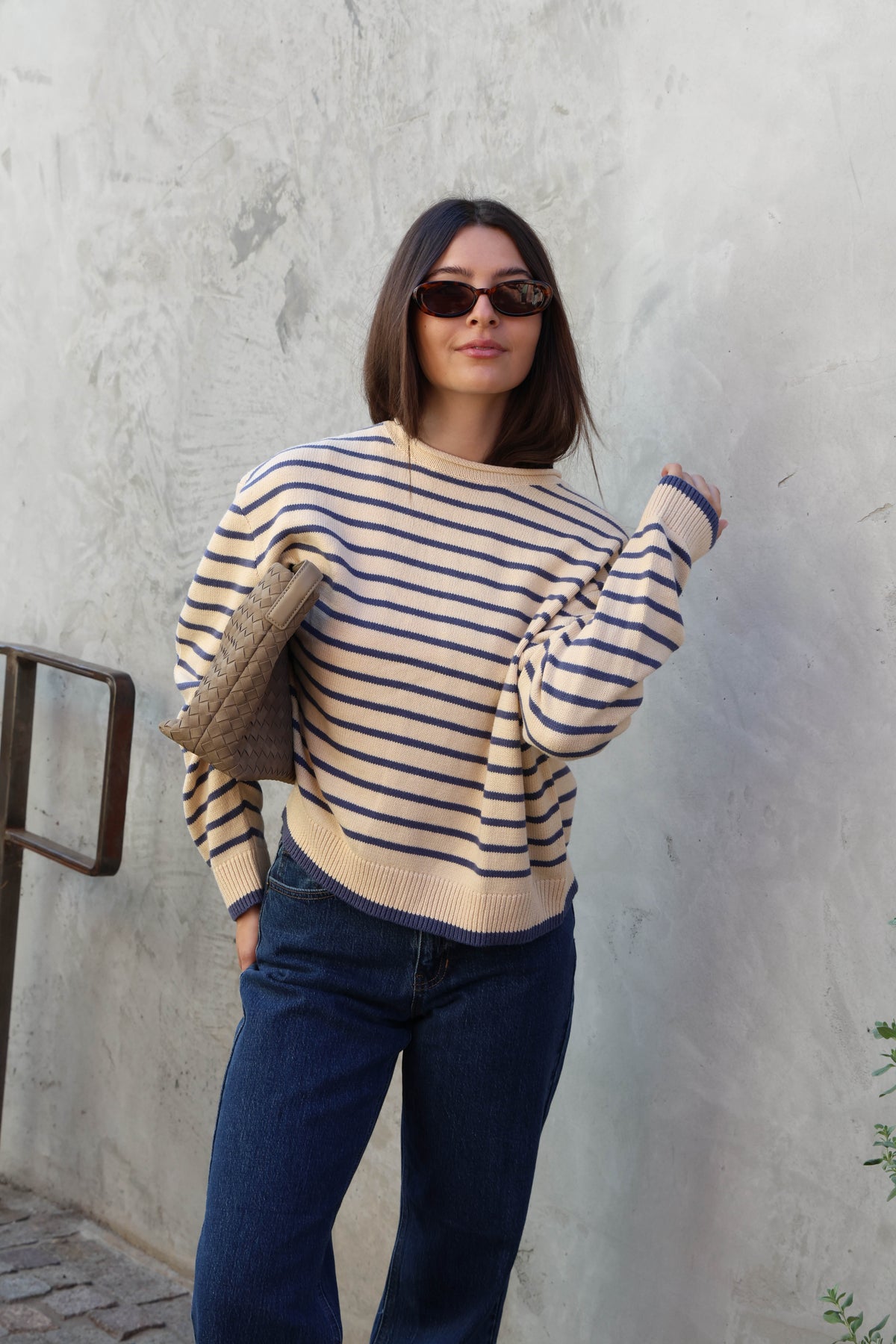 Woman wearing a cream and blue striped sweater with jeans and sunglasses standing against a plain background.