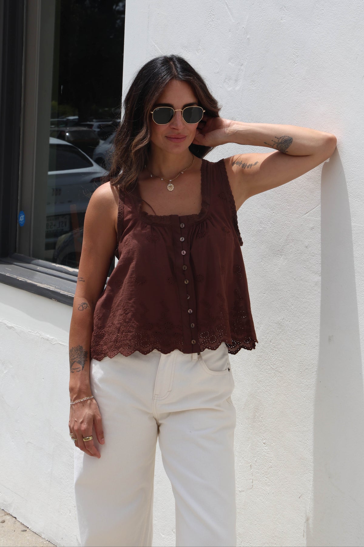 A woman wearing a brown sleeveless lace top and white pants stands against a white stucco wall next to a window.