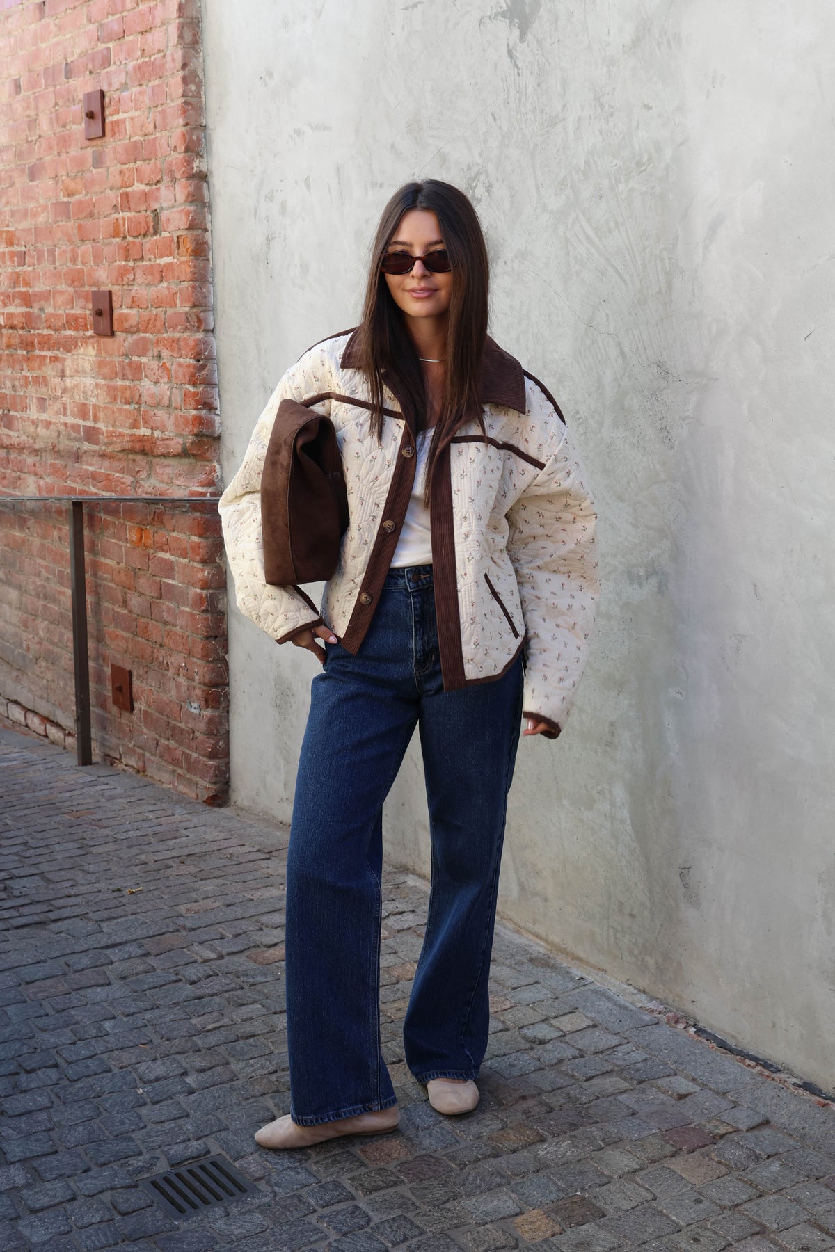 Woman wearing a brown jacket, brown pants holding a brown bad in front of a plain wall.