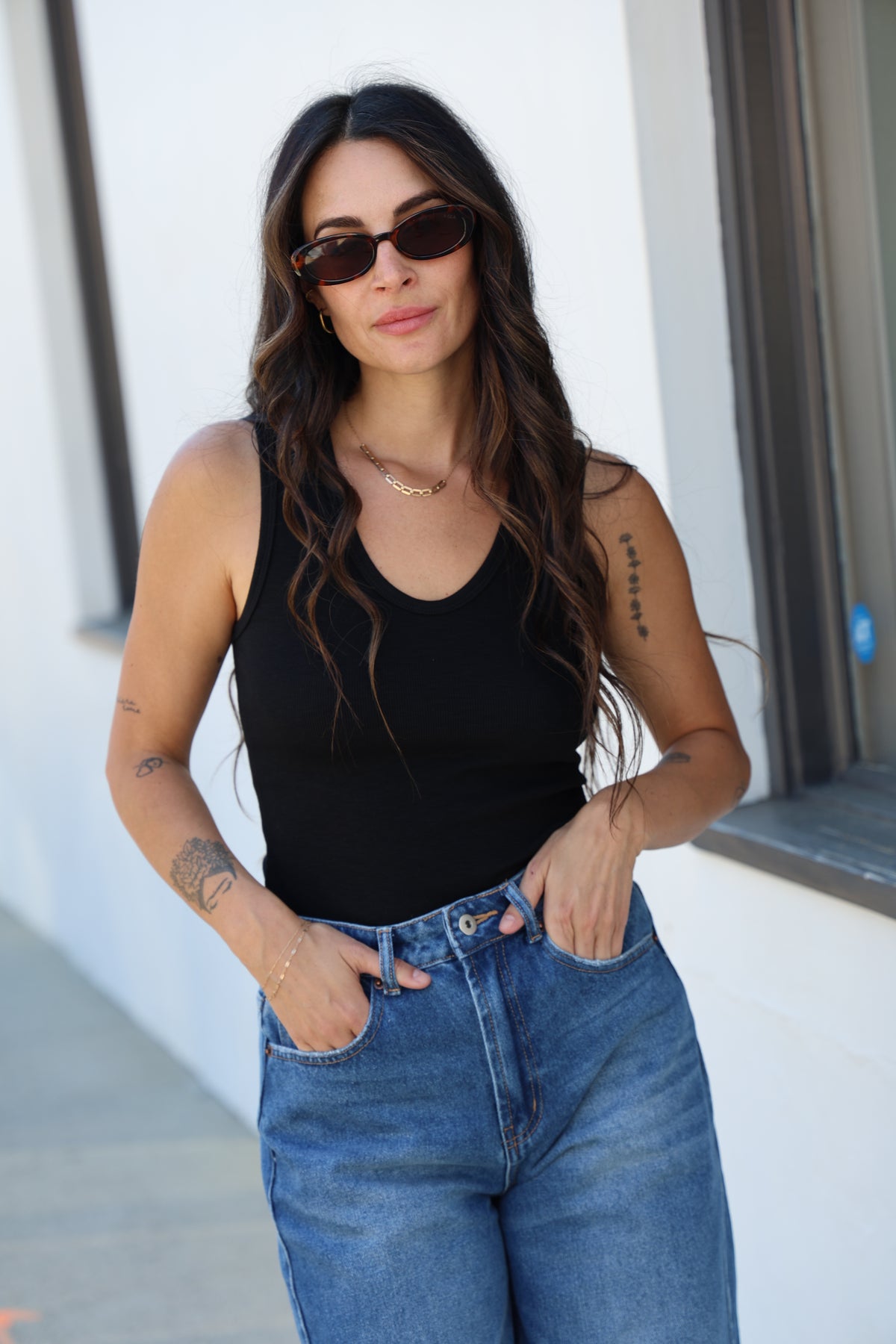 Woman wearing sunglasses and a black top with blue jeans on a white surface.