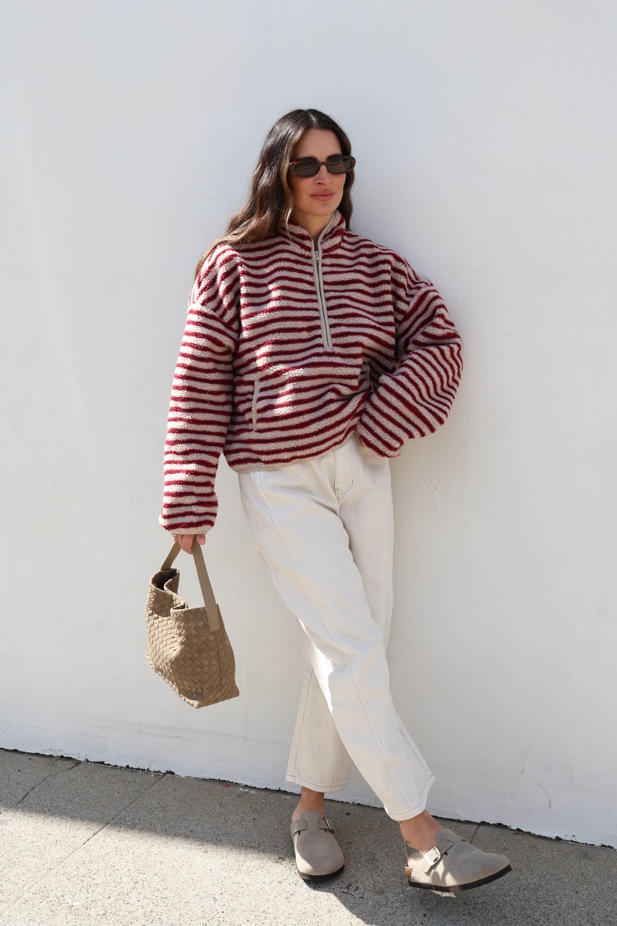 Woman wearing a striped cream and red fleece, sunglasses and white jeans holding a bag.