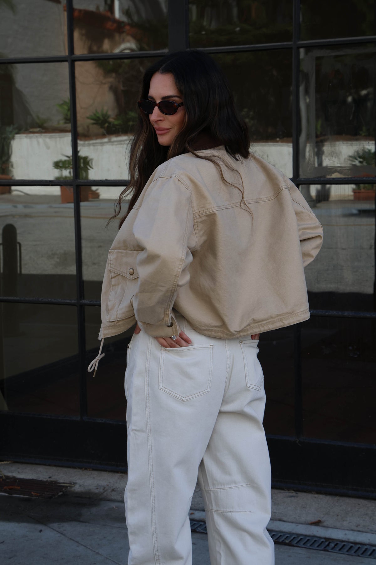 Model is wearing a brown canvas jacket with a white tank, white jeans and sunglasses.