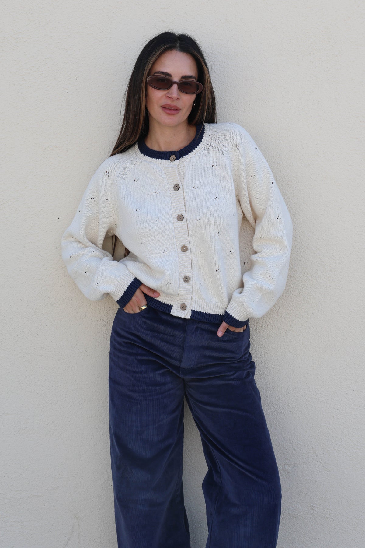 Woman wearing a knit cardigan and blue pants against a plain wall.