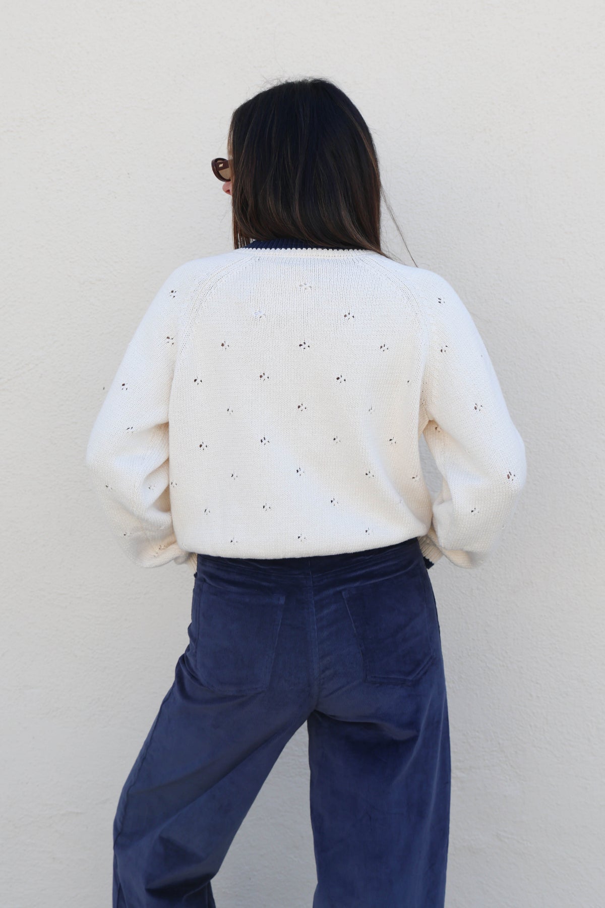 Woman wearing a knit cardigan and blue pants against a plain wall.