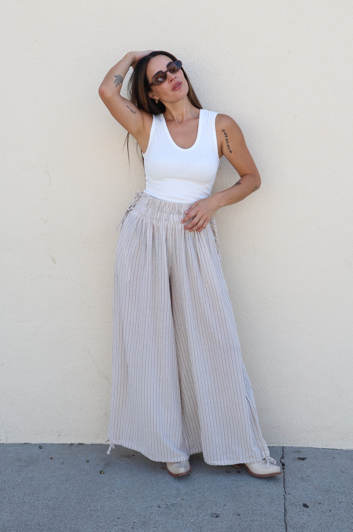 Woman wearing a white tank and striped pants against a plain wall.