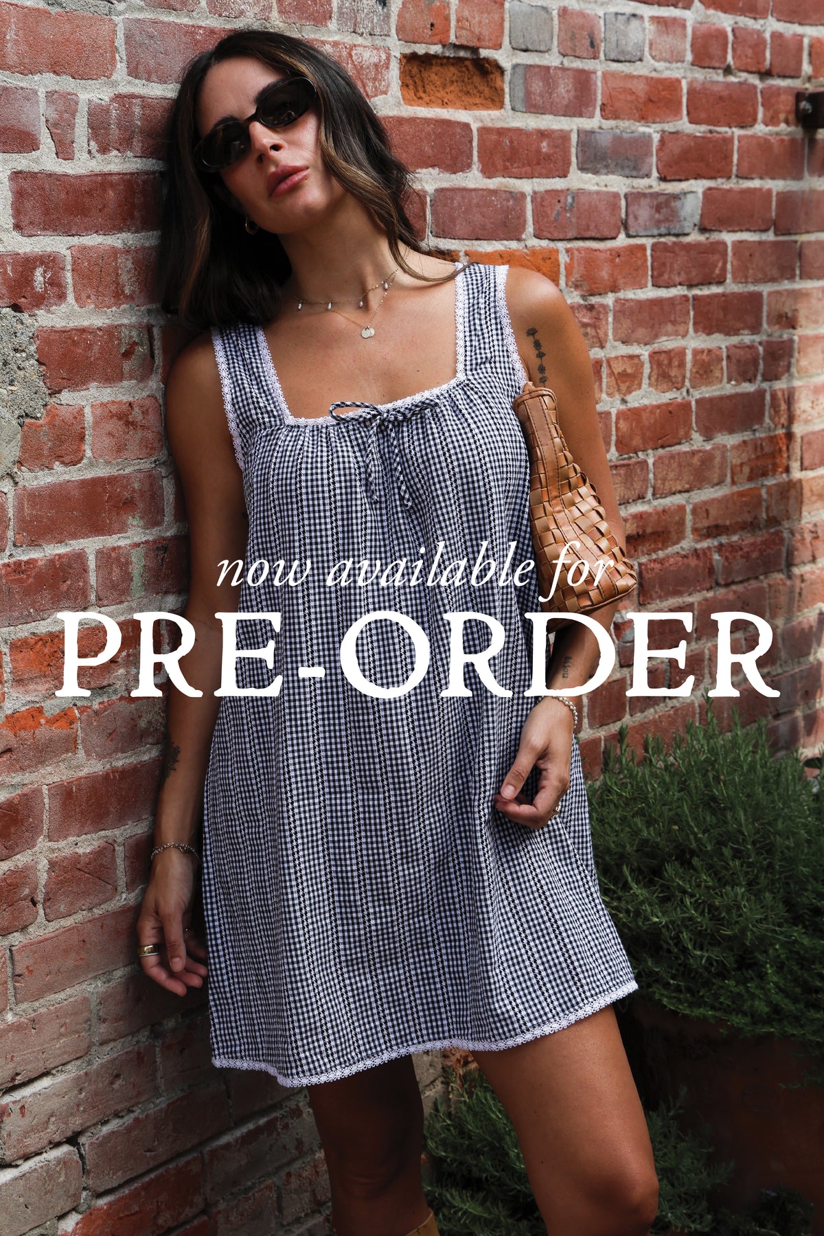 "Now available for Pre-Order" text across the image. Woman wearing a gingham dress against a brick wall.