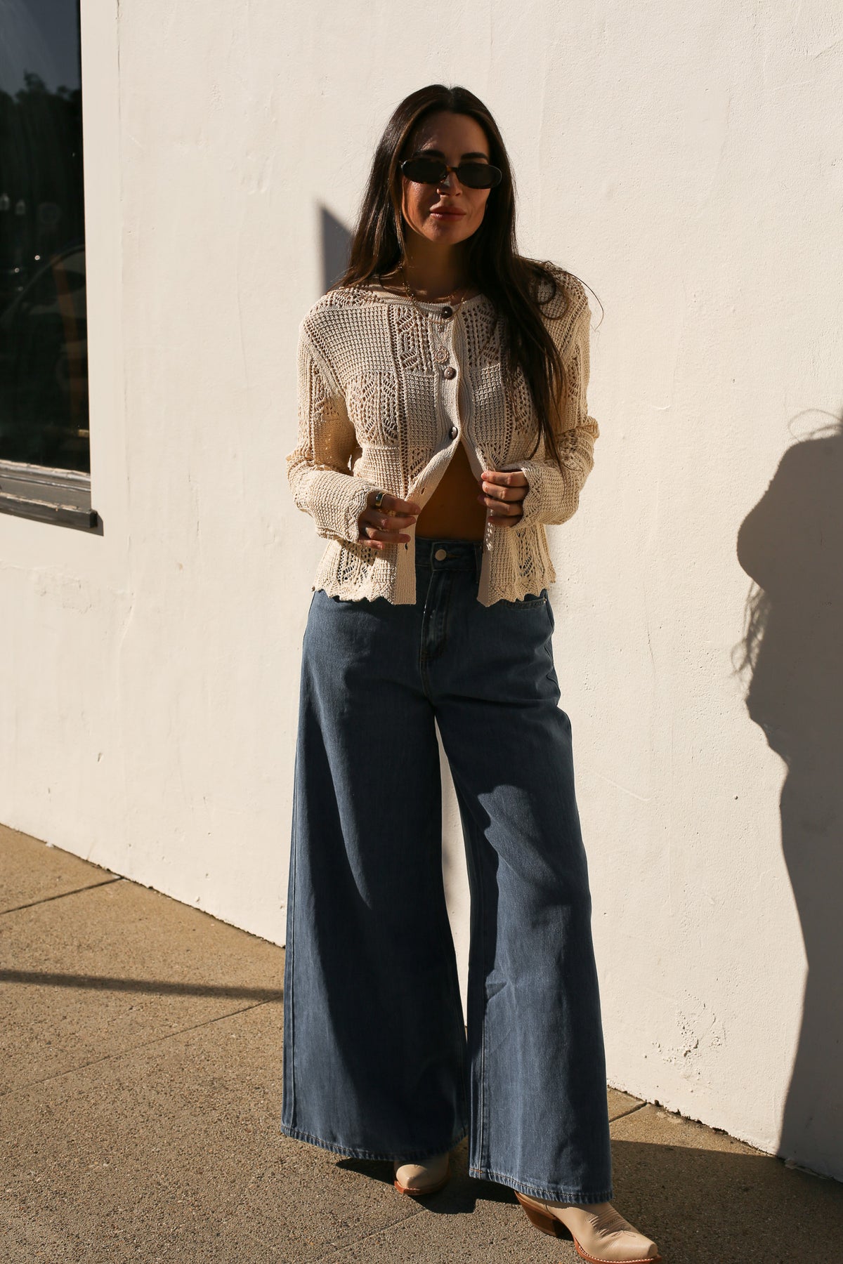 Woman wearing a beige cardigan and blue jeans standing against a white wall.