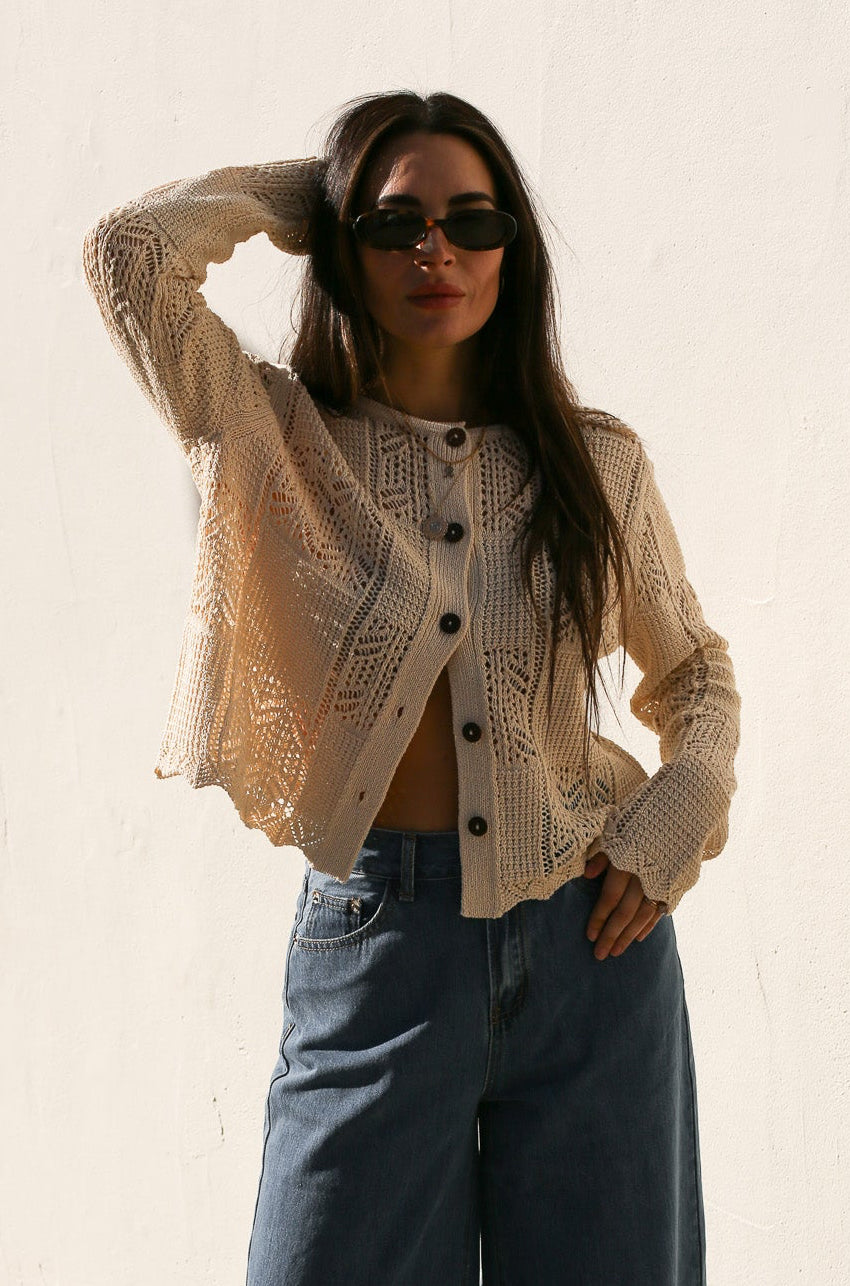 Woman wearing a beige knitted cardigan and sunglasses against a plain background