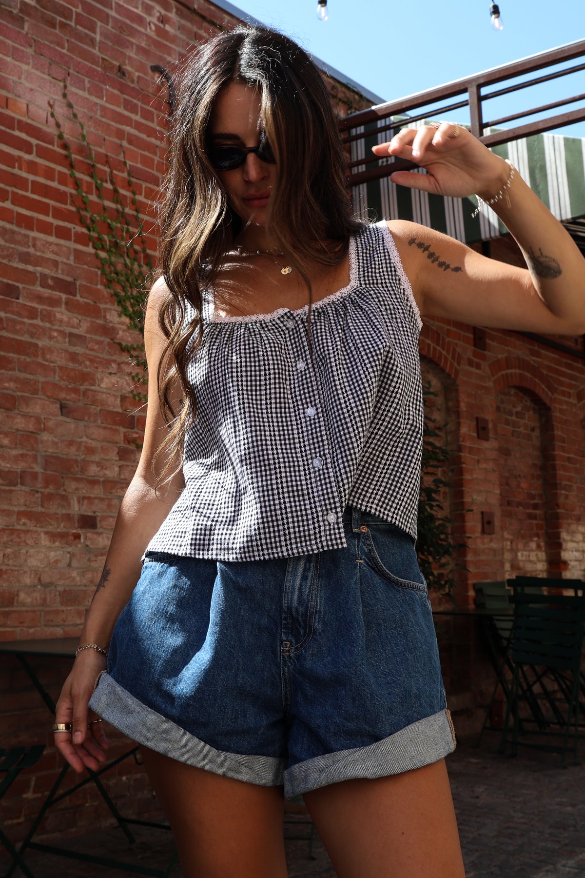 Woman wearing a gingham tank with shorts standing against a brick wall.
