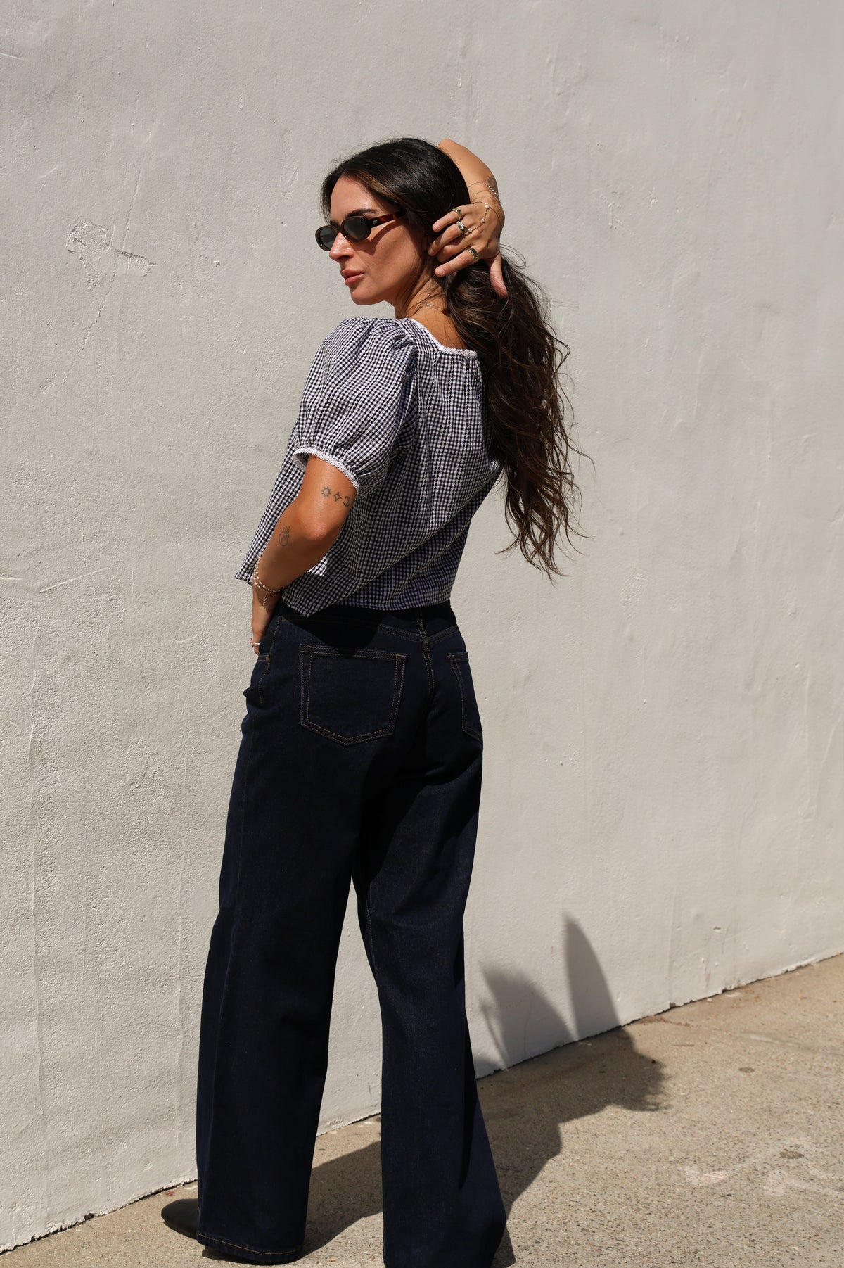Woman wearing a gingham top and jeans against a white wall.