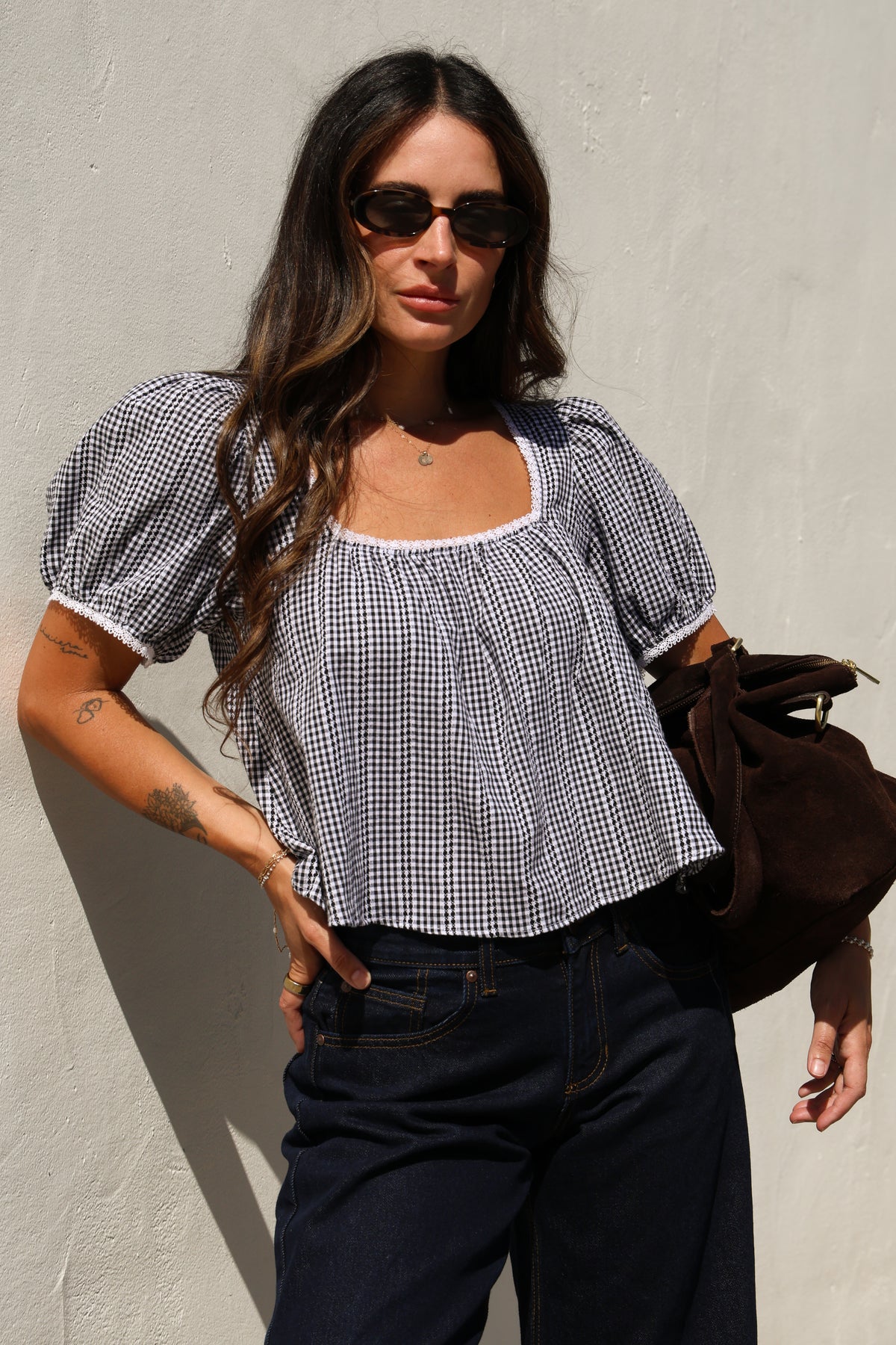 Woman wearing a gingham top and jeans against a white wall.