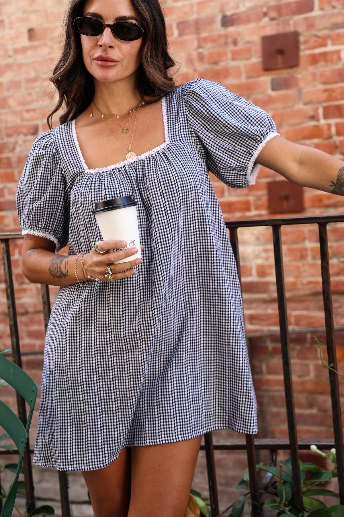Woman wearing a gingham dress with sunglasses standing holding a coffee cup.