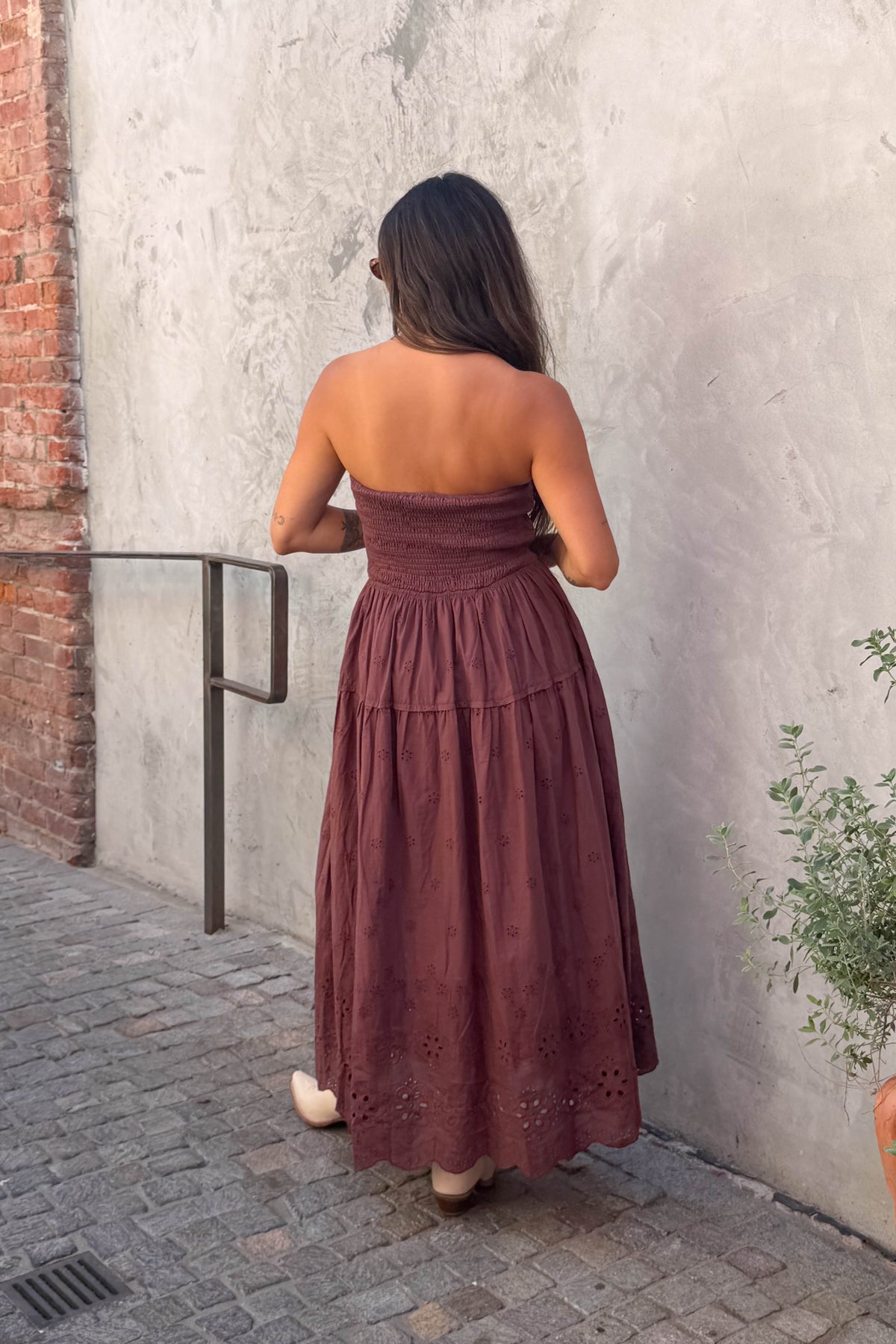 A woman wearing a strapless rust-brown tiered midi dress with eyelet lace details and nude heels, posing against a textured concrete wall.
