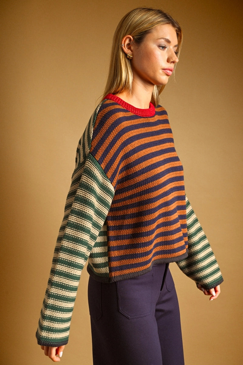 A woman wearing a striped knit sweater with a navy and rust-orange striped body and green-and-beige striped sleeves, paired with dark trousers, set against a solid warm brown background.