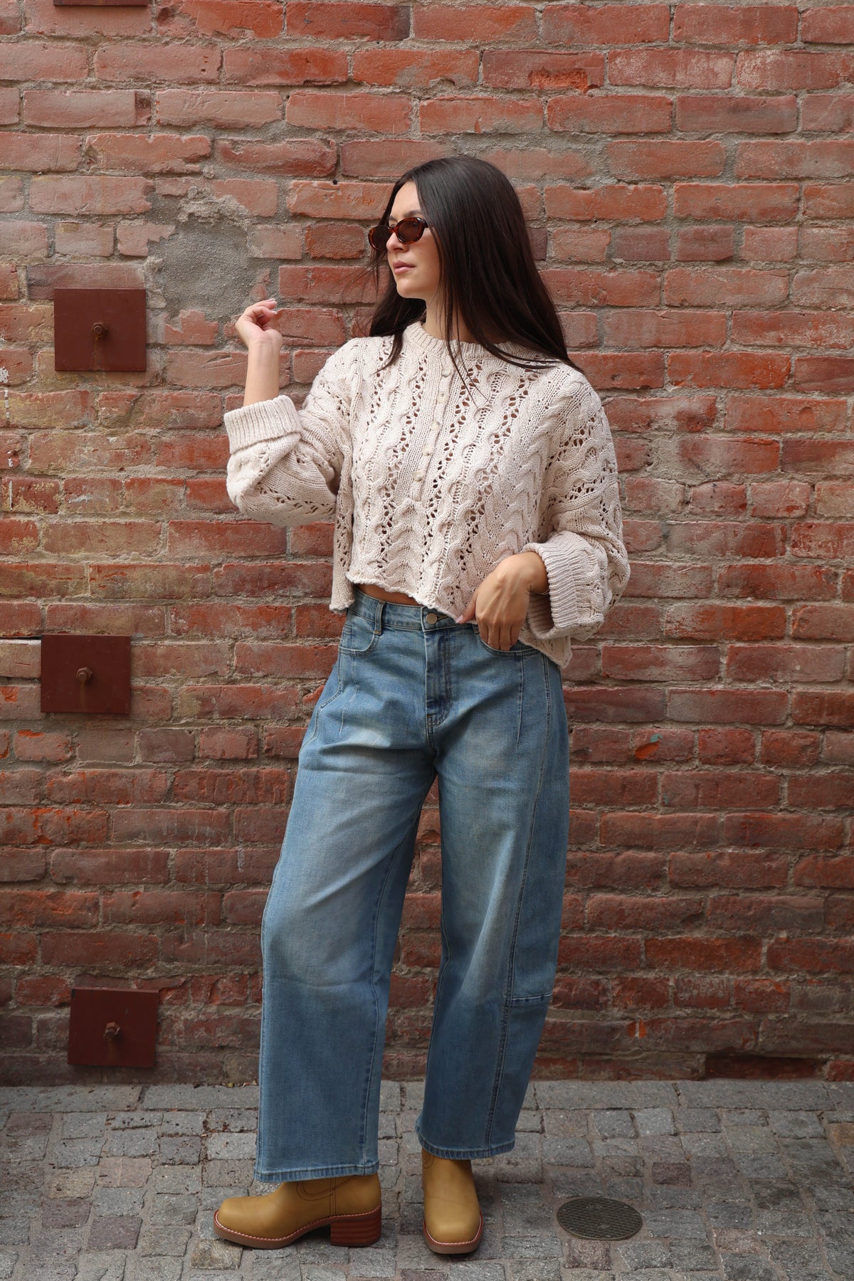 A woman stands against a red brick wall wearing a cream-colored crochet knit sweater, wide-leg blue jeans, and tan ankle boots.