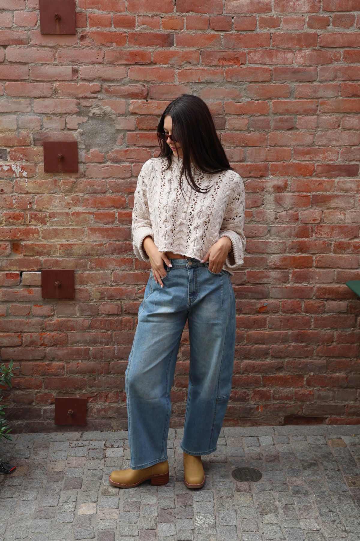 Woman wearing barrel jeans with a cream knit against a brick wall.
