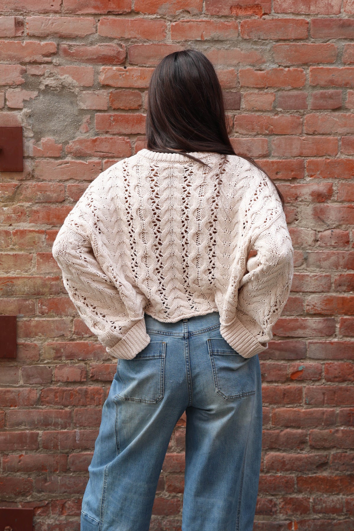 Woman wearing a crochet sweater in oatmeal with jeans against a brick wall.