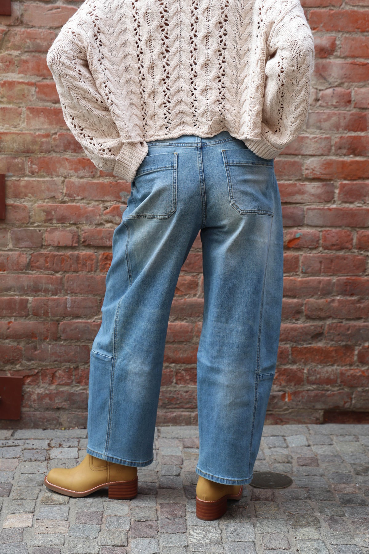 Woman wearing barrel jeans with a cream knit against a brick wall.