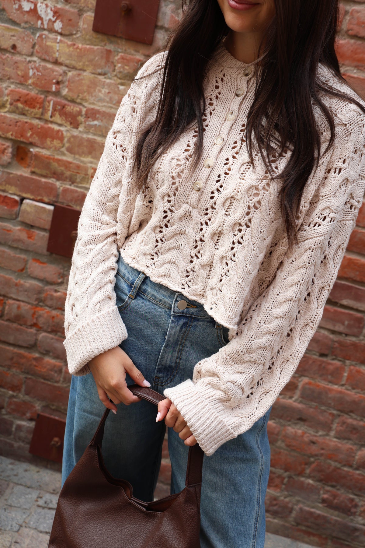 A woman stands against a red brick wall wearing a cream-colored crochet knit sweater, wide-leg blue jeans, and tan ankle boots.