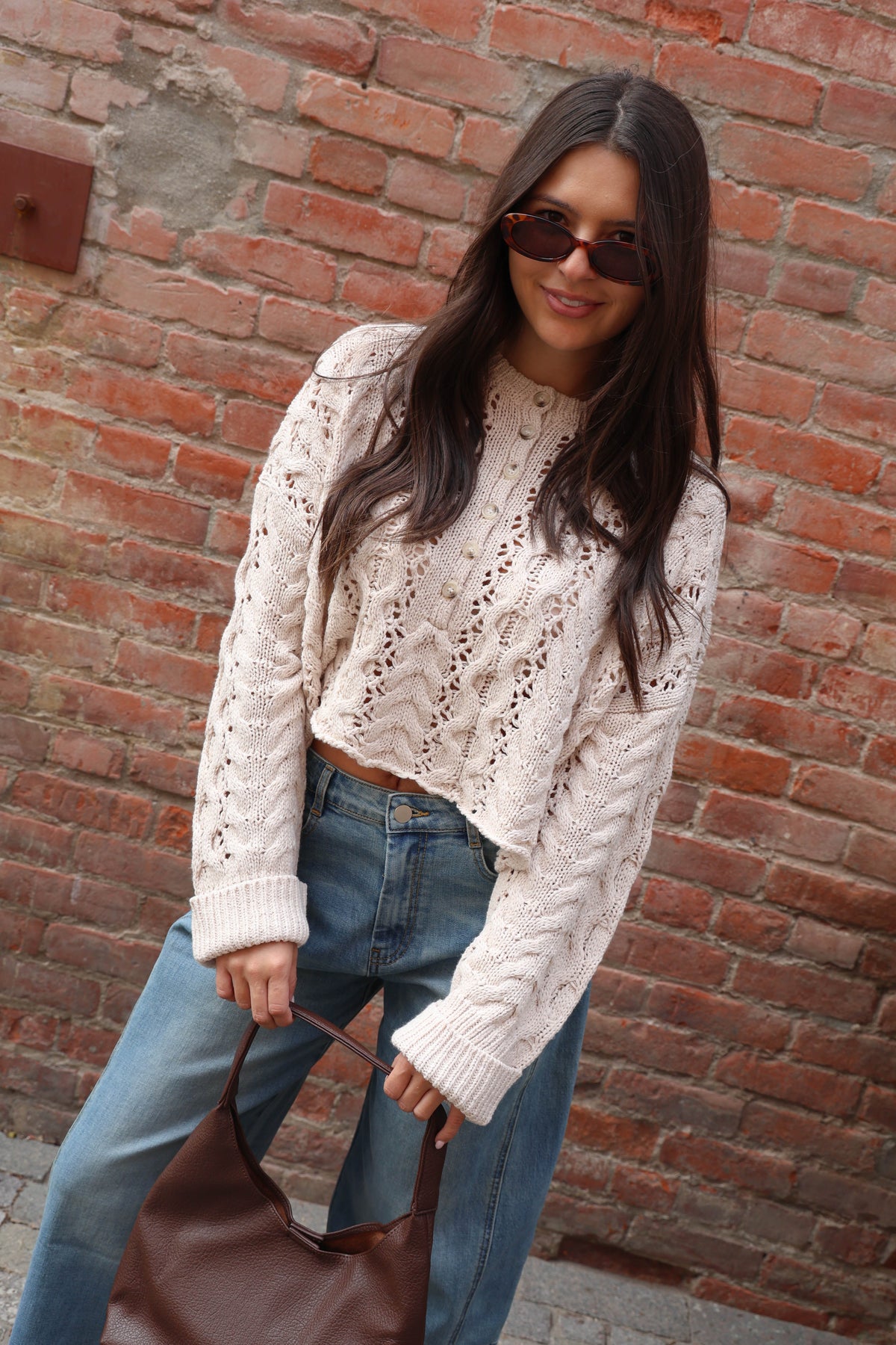 Woman wearing a crochet sweater in oatmeal with jeans against a brick wall.