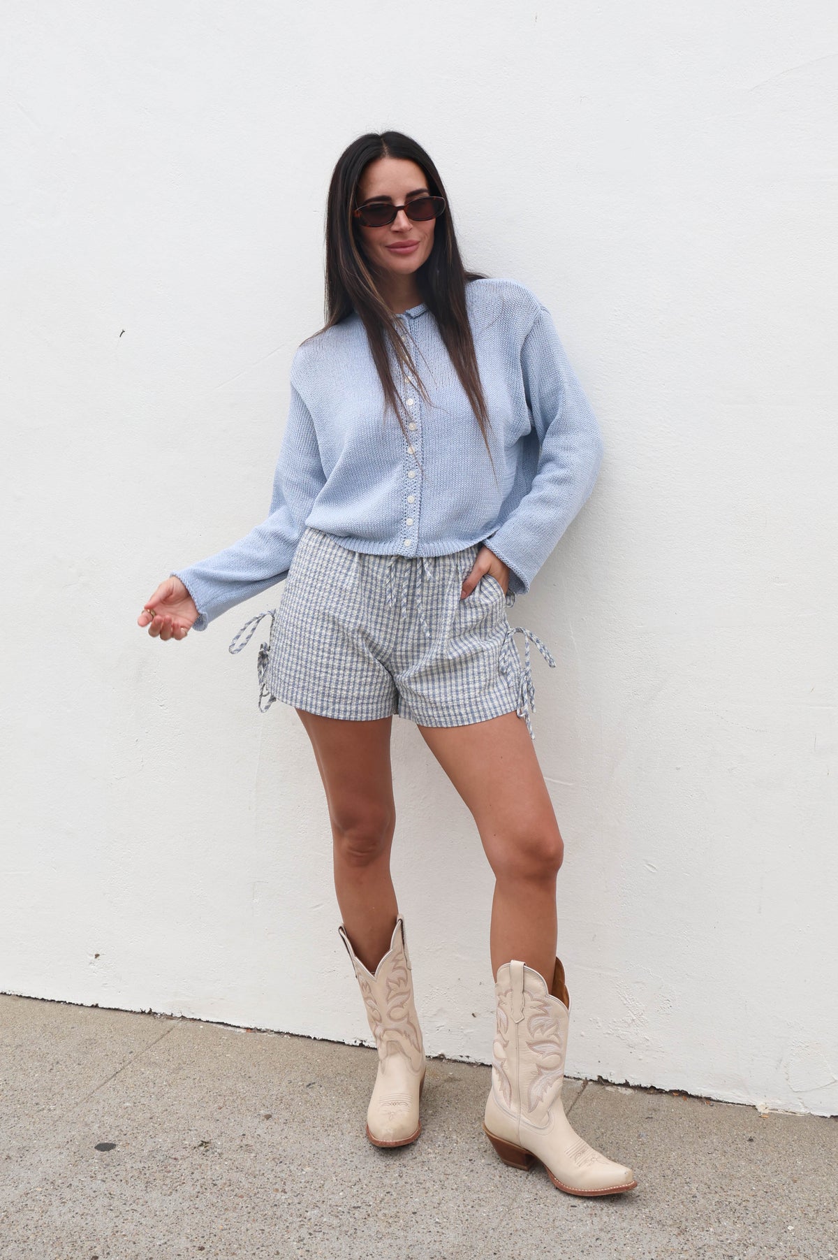 Woman wearing a light blue shirt, checkered shorts, and beige boots against a white wall.