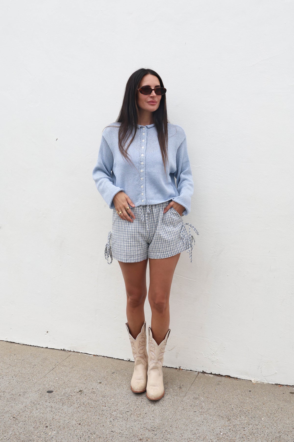 Woman wearing a light blue shirt, checkered shorts, and white boots against a white wall.