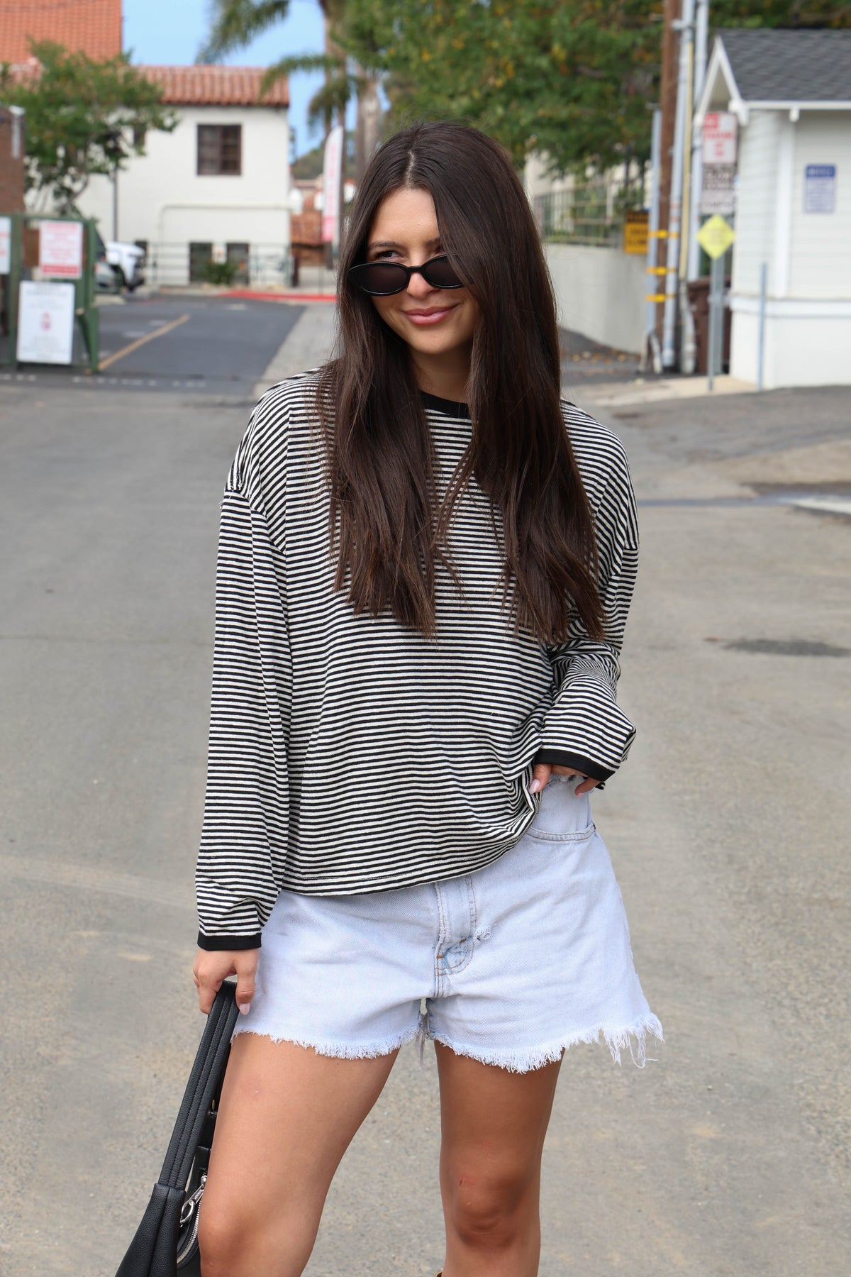 A woman wearing a black and white striped long-sleeve t-shirt and light wash denim shorts, standing on an outdoor street.