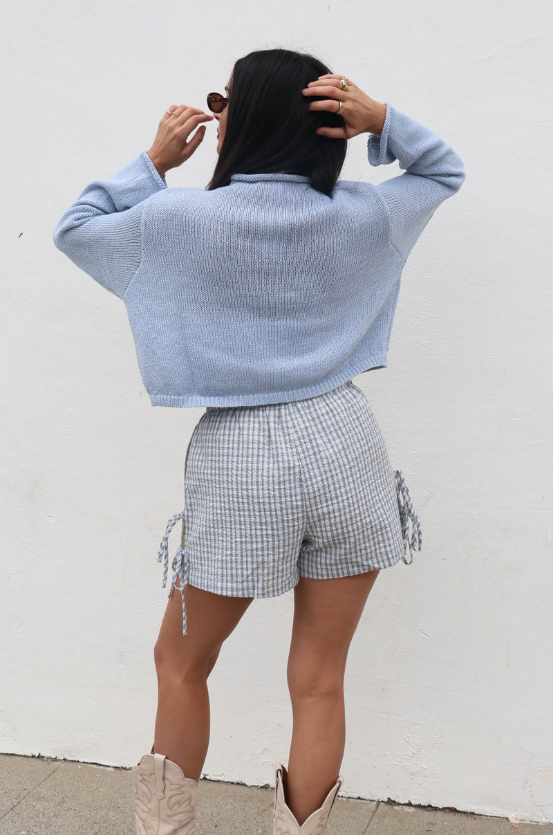 Person wearing a light blue sweater, checkered shorts, and beige boots against a white wall.