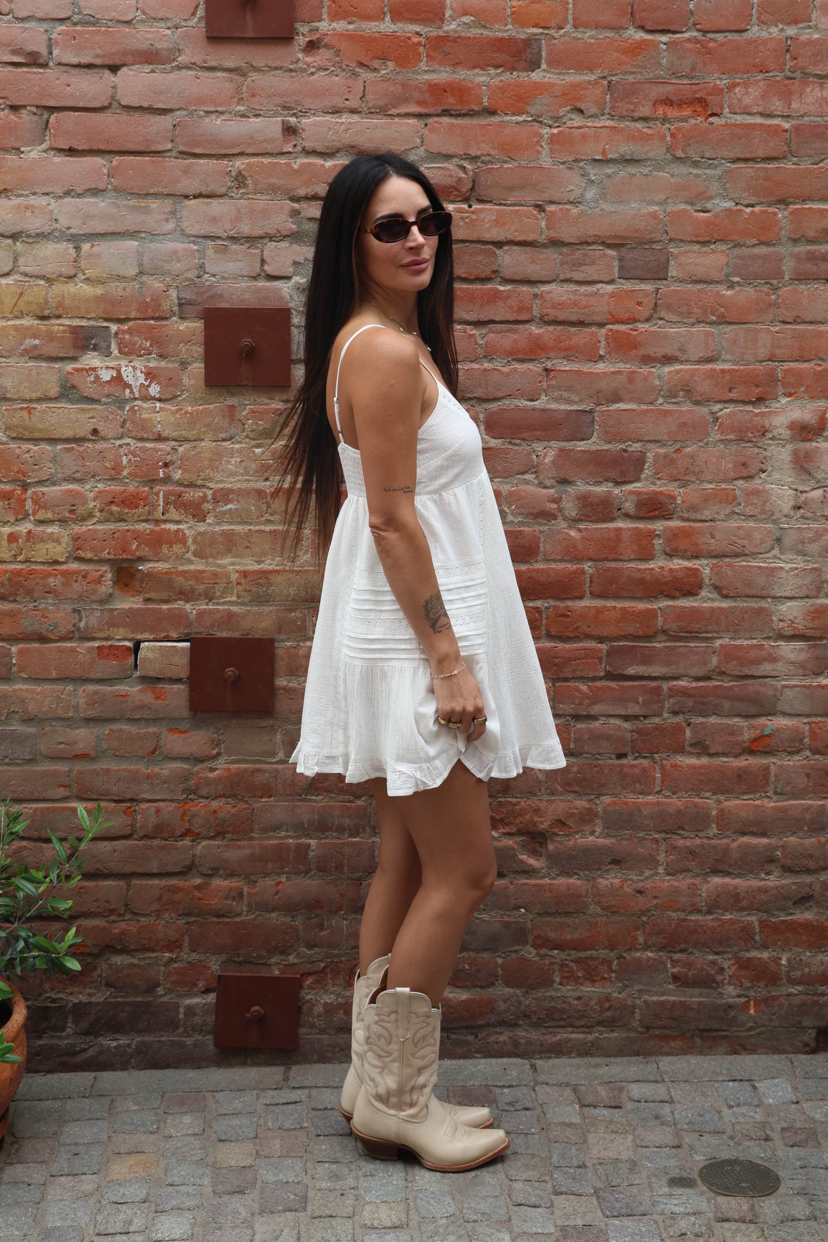 Woman in a white dress and beige boots standing against a brick wall.