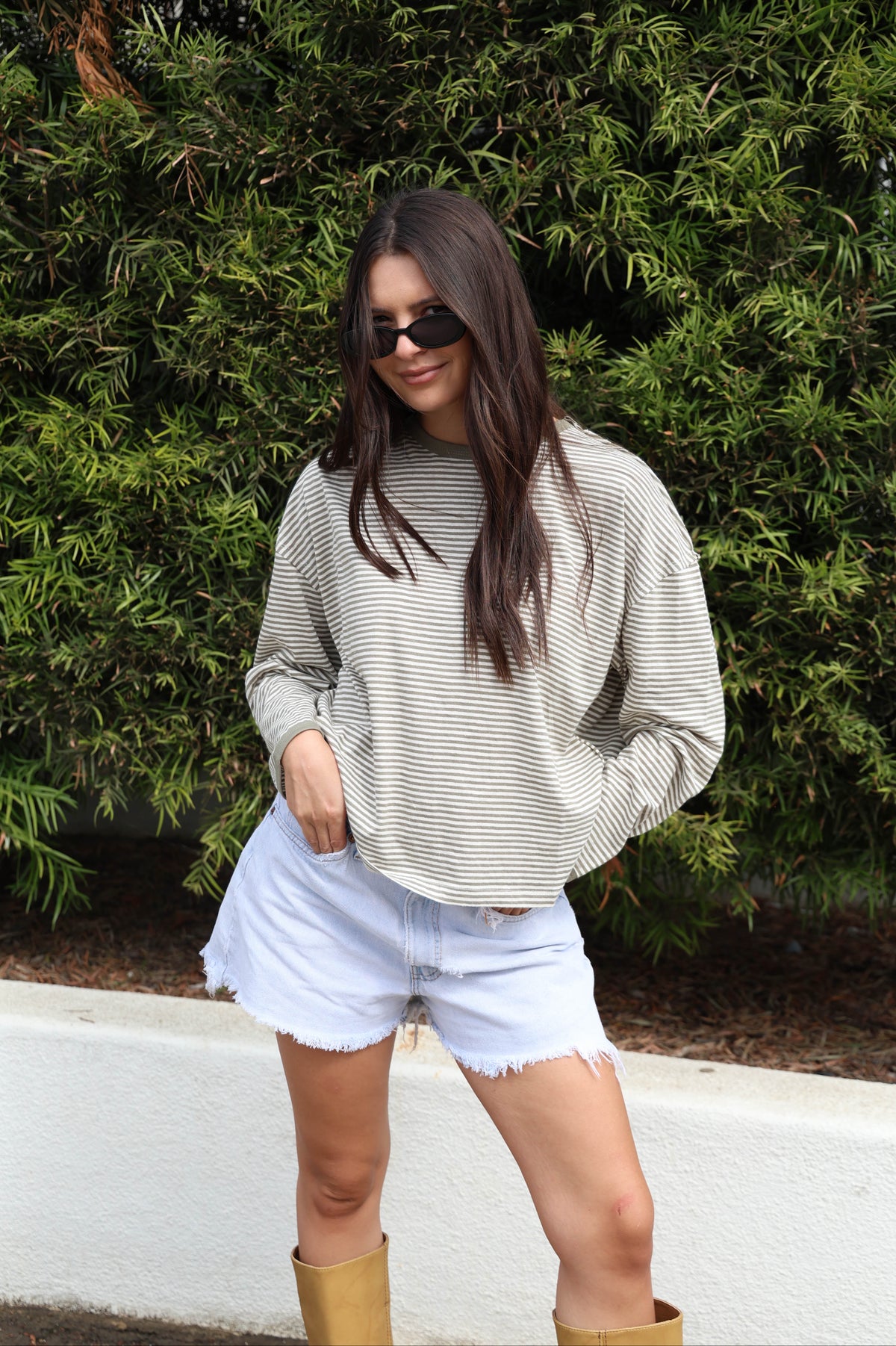 Woman wearing a striped long sleeve shirt with jean shorts and boots against a bush.