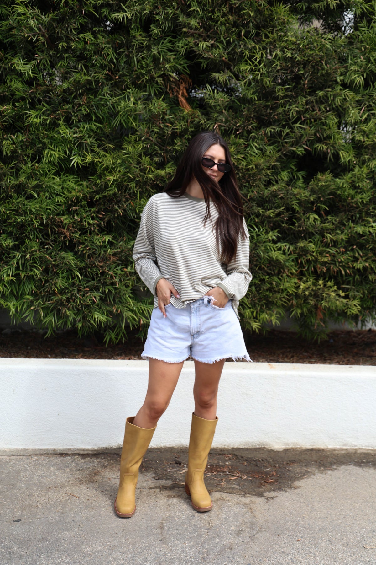 Woman wearing a striped long sleeve shirt with jean shorts and boots against a bush.