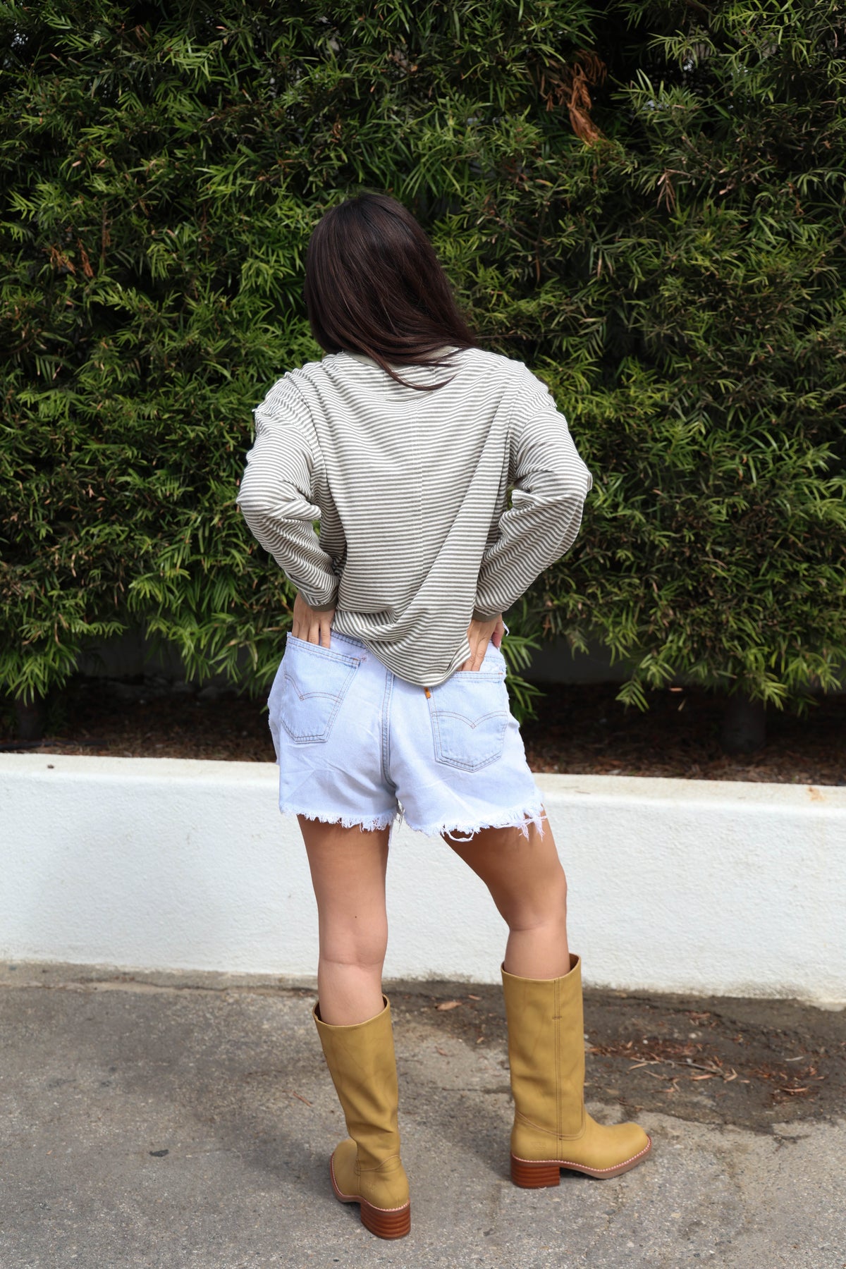 Woman wearing a striped long sleeve shirt with jean shorts and boots against a bush.