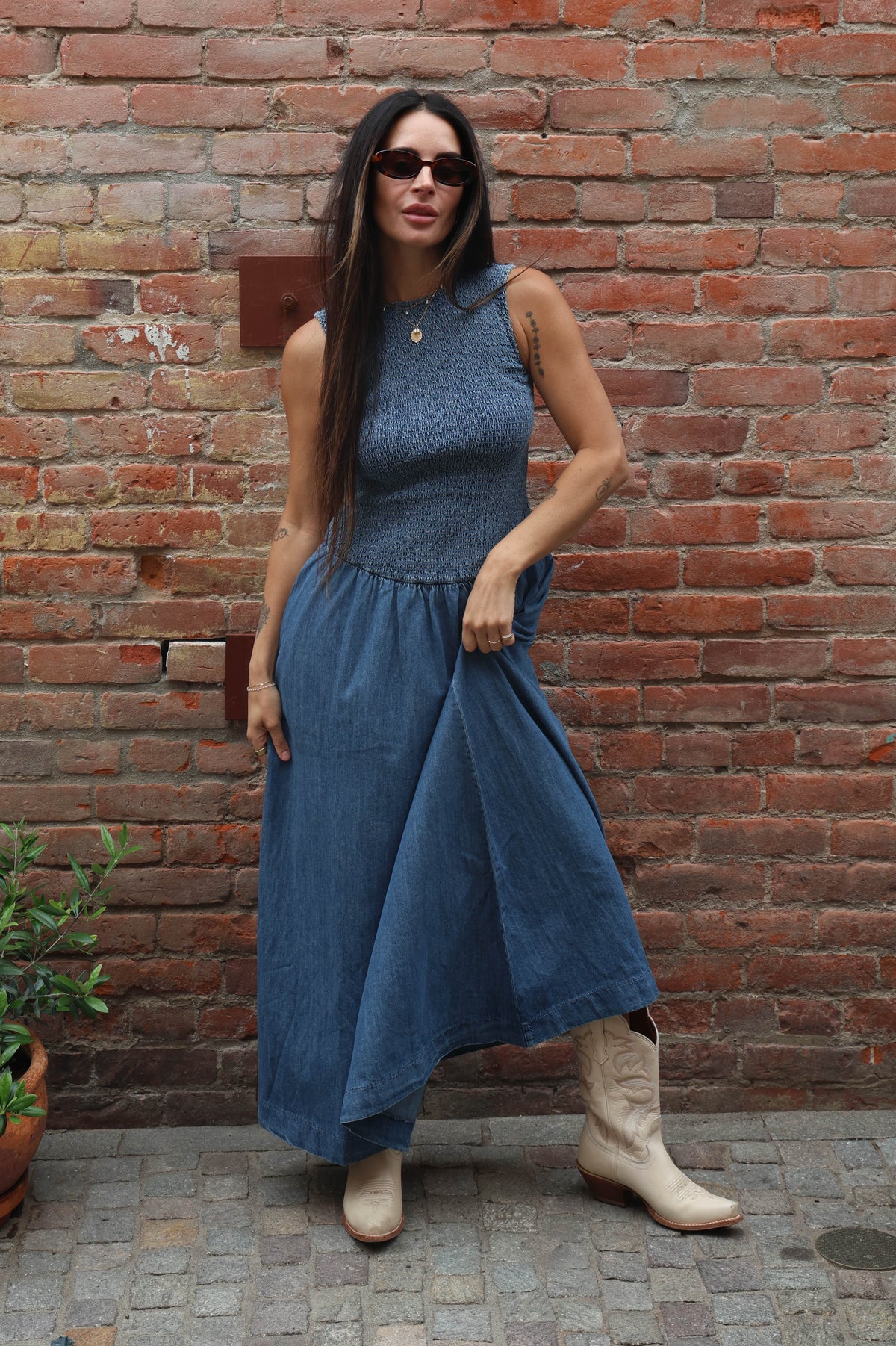 Woman in a blue dress and beige boots standing against a brick wall.