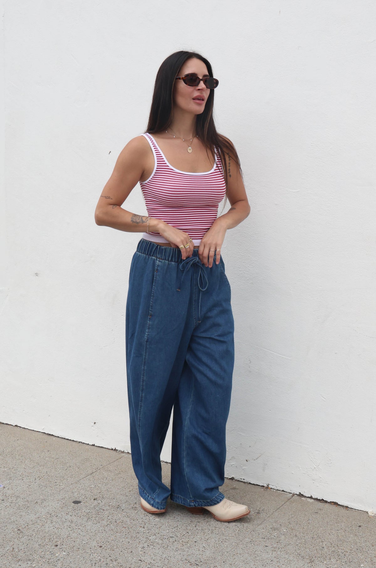 Woman wearing a striped tank top and blue jeans standing against a white wall.